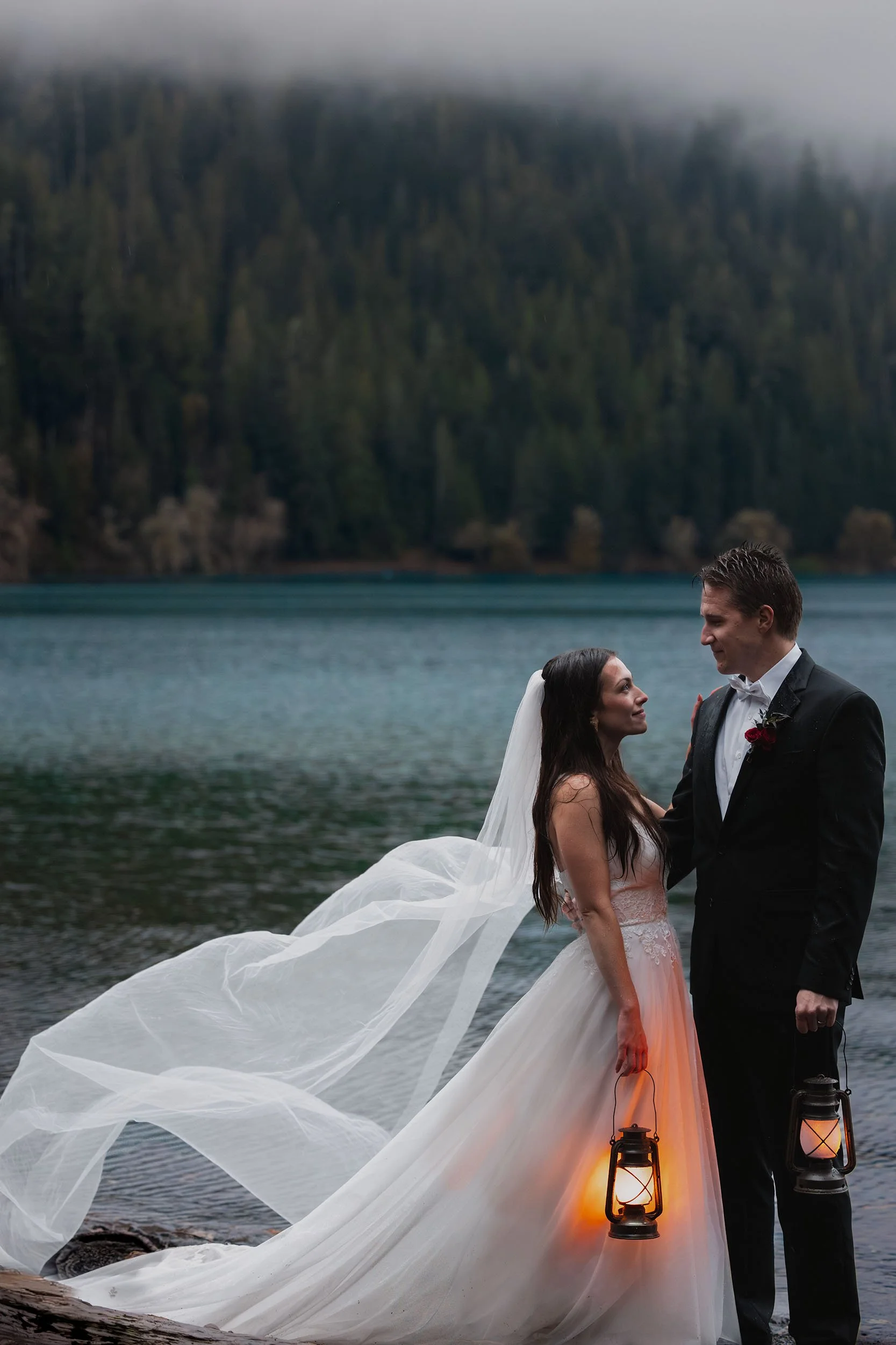 A couple in wedding attire hold lanterns in the rain at Lake Crescent. The bride's veil blows dramatically in the wind.