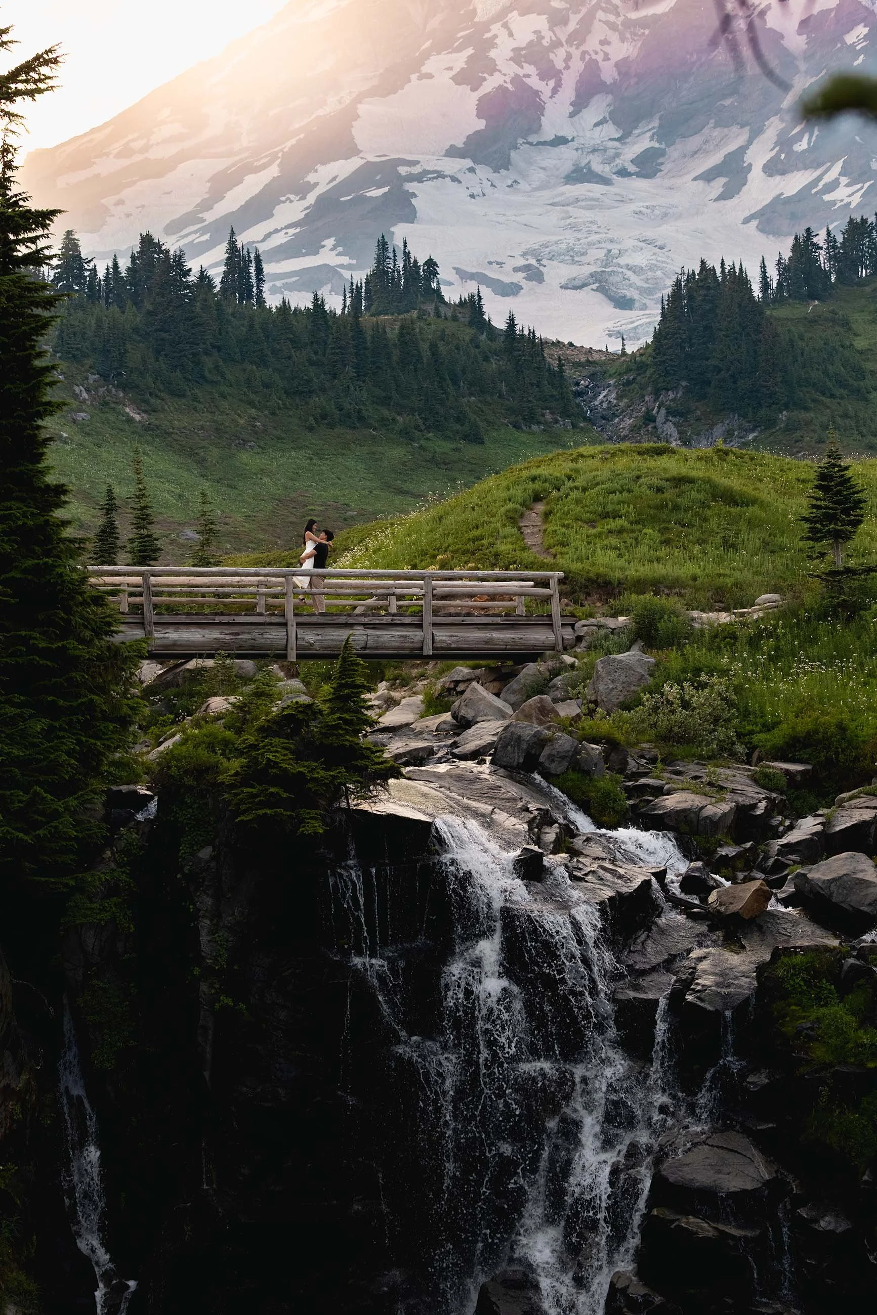 A man lifts his fiance on the bridge over Myrtle Falls during a wildflower engagement session at sunset. Mount Rainier looms large in the background.