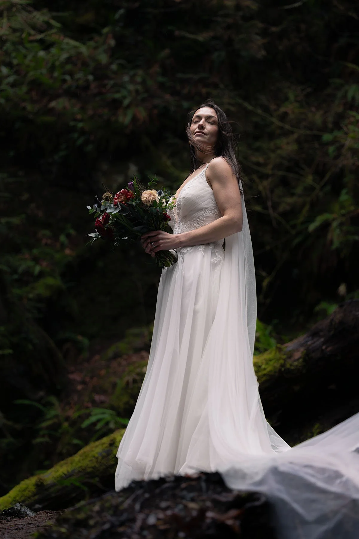 A bride standing in a dark mossy forest at Olympic National Park holds a bouquet and the wind blows her hair.