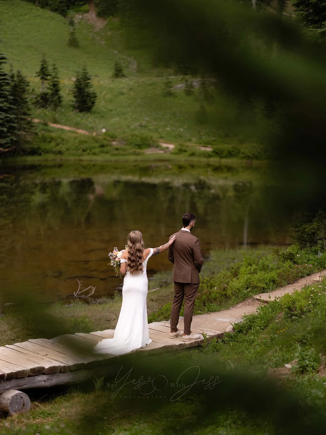A bride in a white gown taps her fiance on the shoulder on a wood bridge by a lake for their first looks on their wedding day.