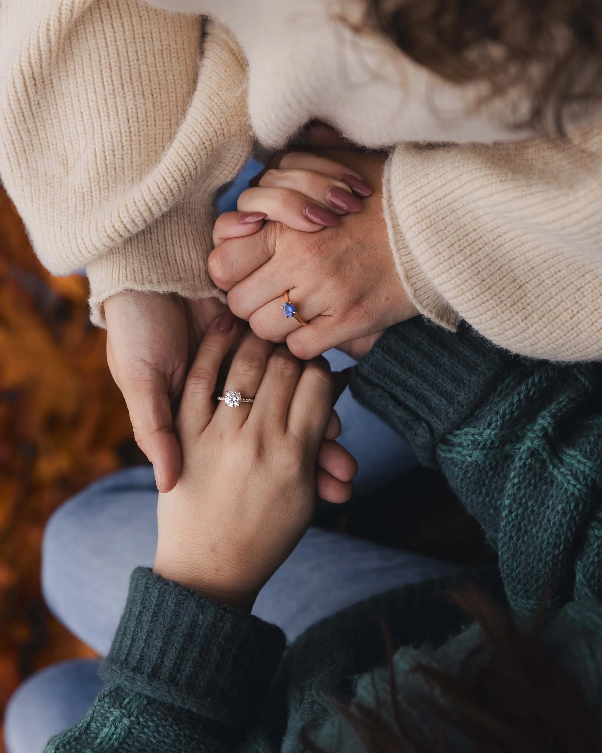 LGBTQ couple hold hands showing off their engagement rings. Orange fall leaves are on the ground. 