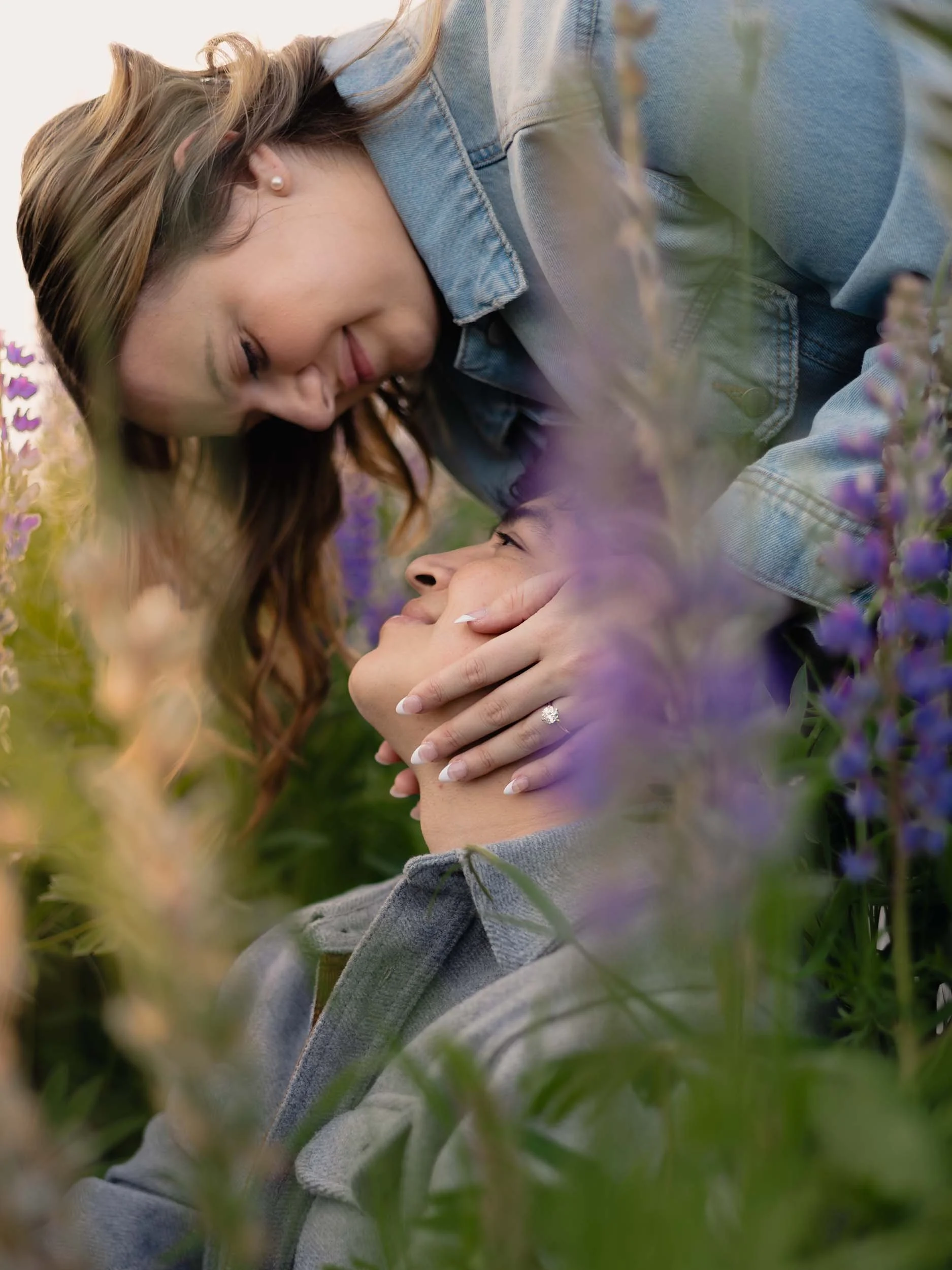 A smiling woman leans over her fiance showing off her engagement ring in a field of purple lupine in Puyallup. 