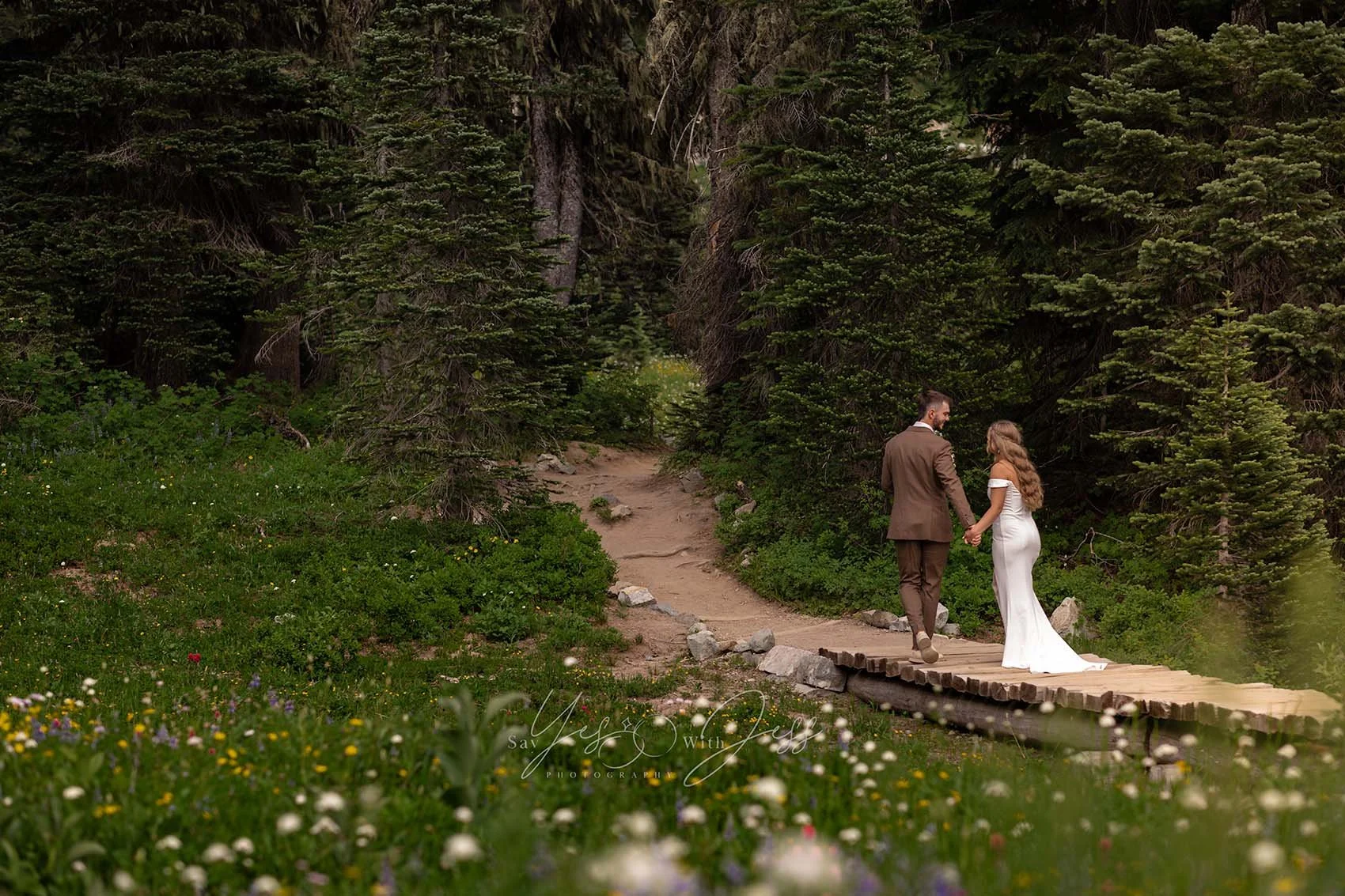 A couple walk across a primitive wooden bridge through a meadow of subalpine wildflowers on their wedding day at Tipsoo Lake.