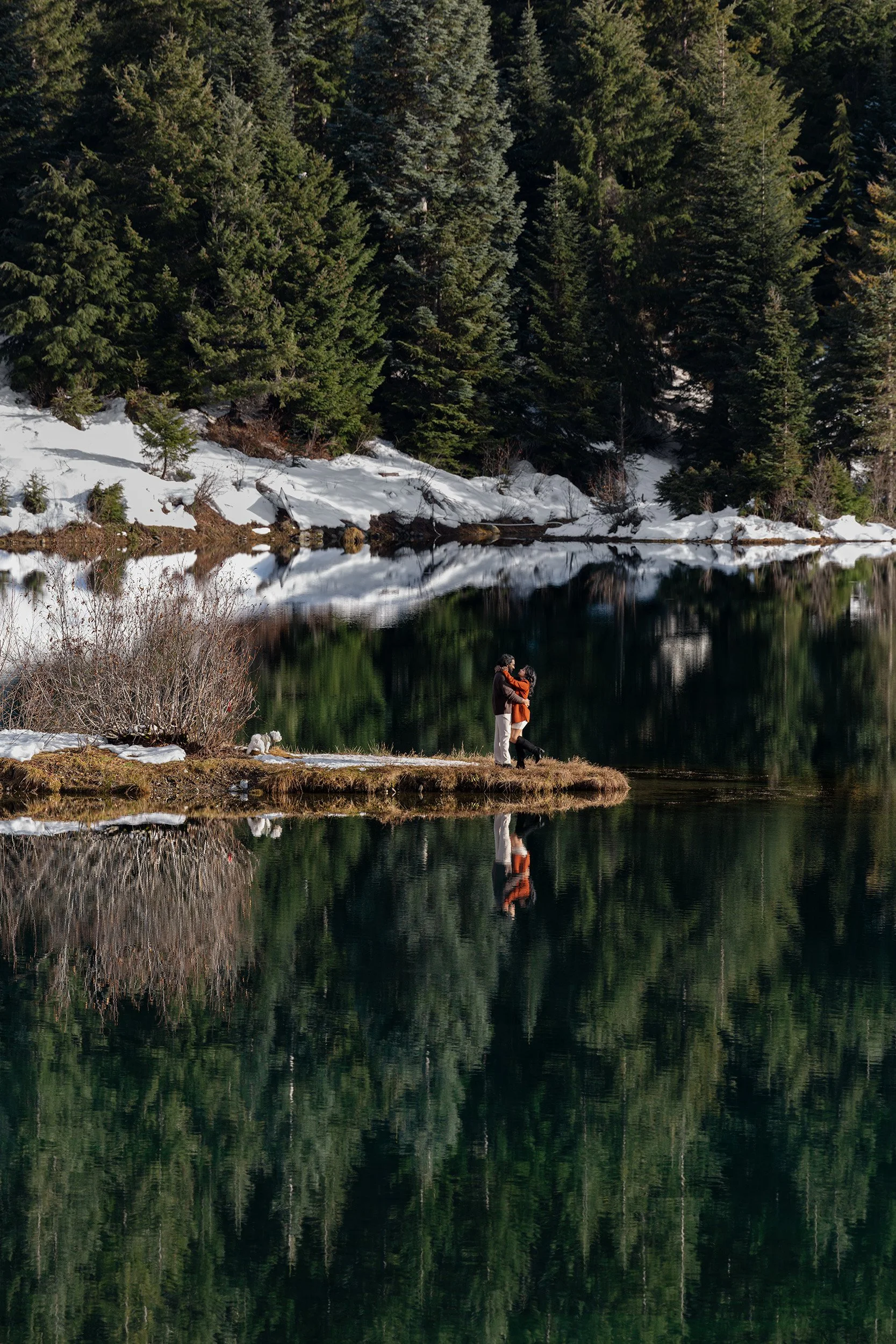 A couple stand on a peninsula jutting into Gold Creek Pond in the winter. The trees and snow are perfectly reflected in the pond like glass.