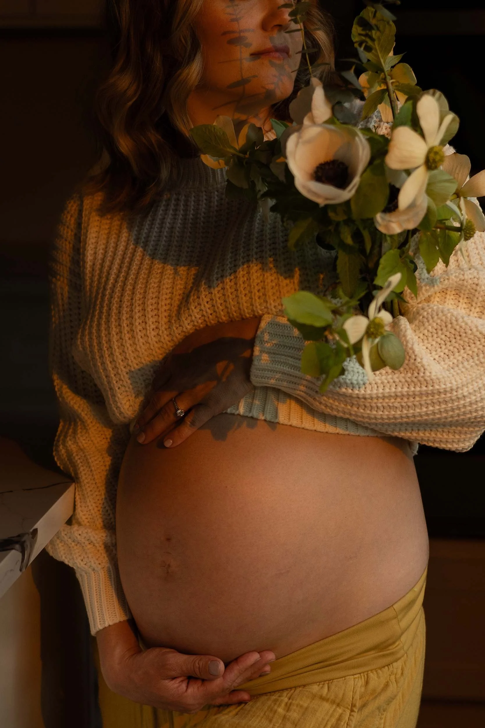 A woman holds a bouquet and her baby bump, with the sunset casting an orange hue, during a lifestyle maternity session near Olympia.