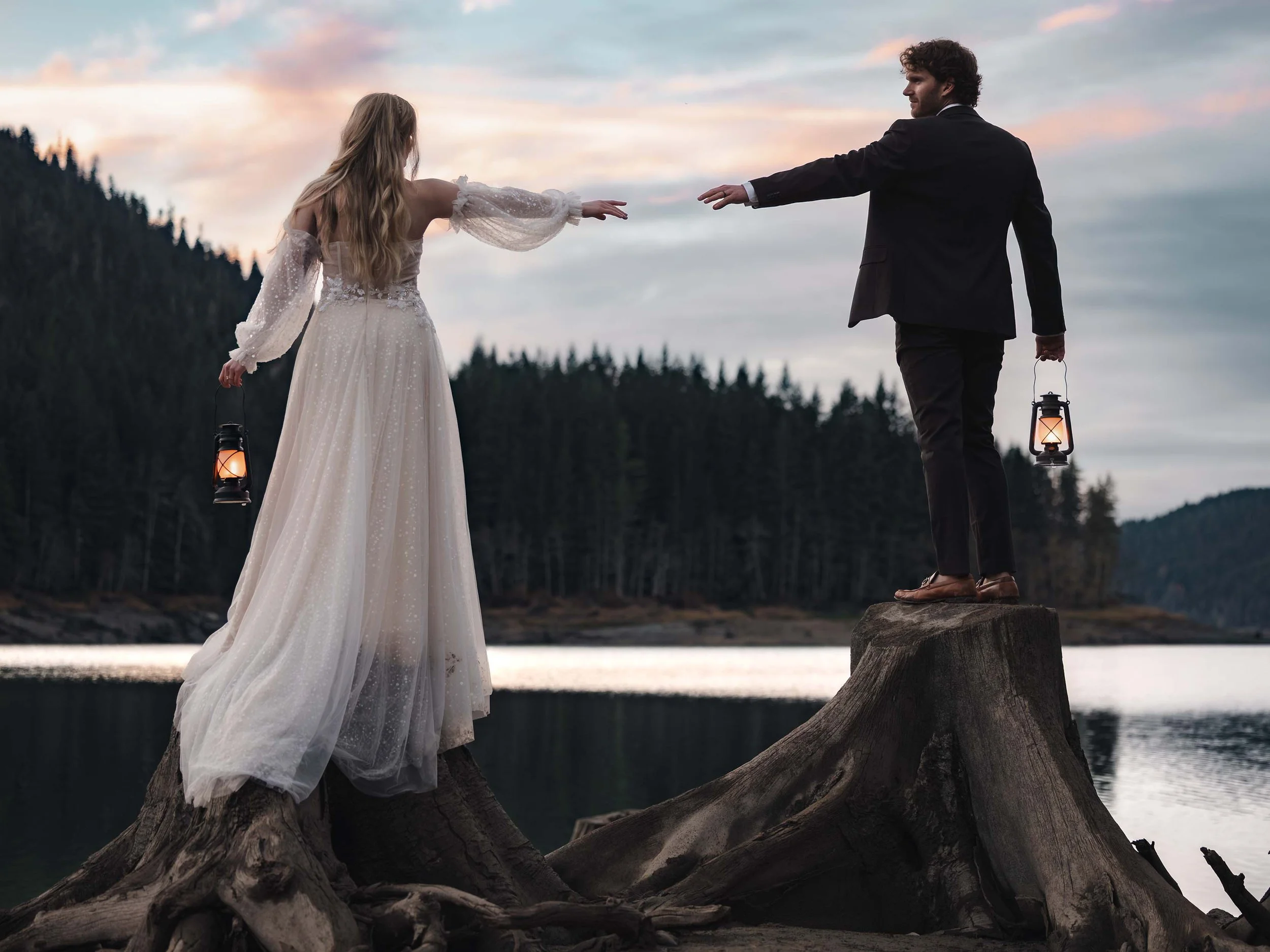 Micro wedding couple holding lanterns reach for each other. They stand on old growth tree stumps at the edge of Greider Lake in Washington.