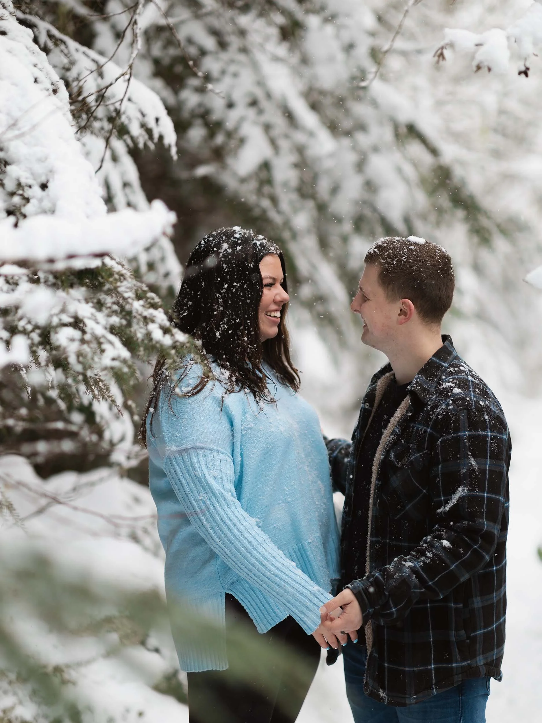 A couple hold hands as they are covered with falling snow at an engagement session in Mount Baker Snoqualmie National Forest.