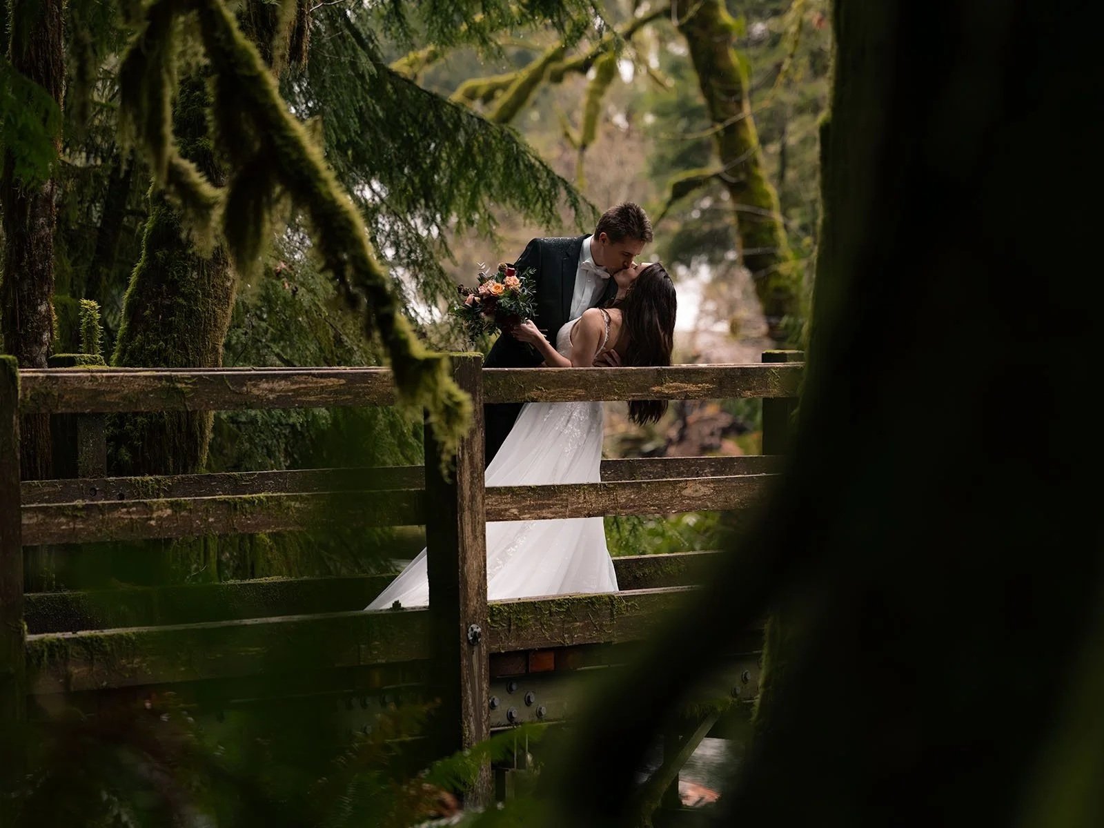 A man in a suit dips his bride holding a bouquet on a bridge surrounded by lush, moss covered trees in Bovees Meadow.