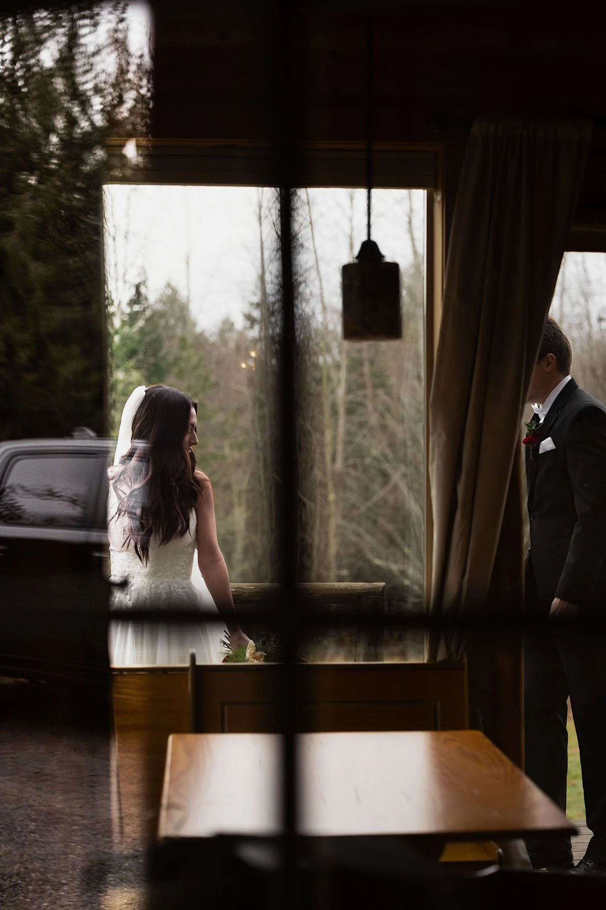 A bride and groom walk on a deck as photographed from inside through a door with a reflection of his truck on their wedding day.