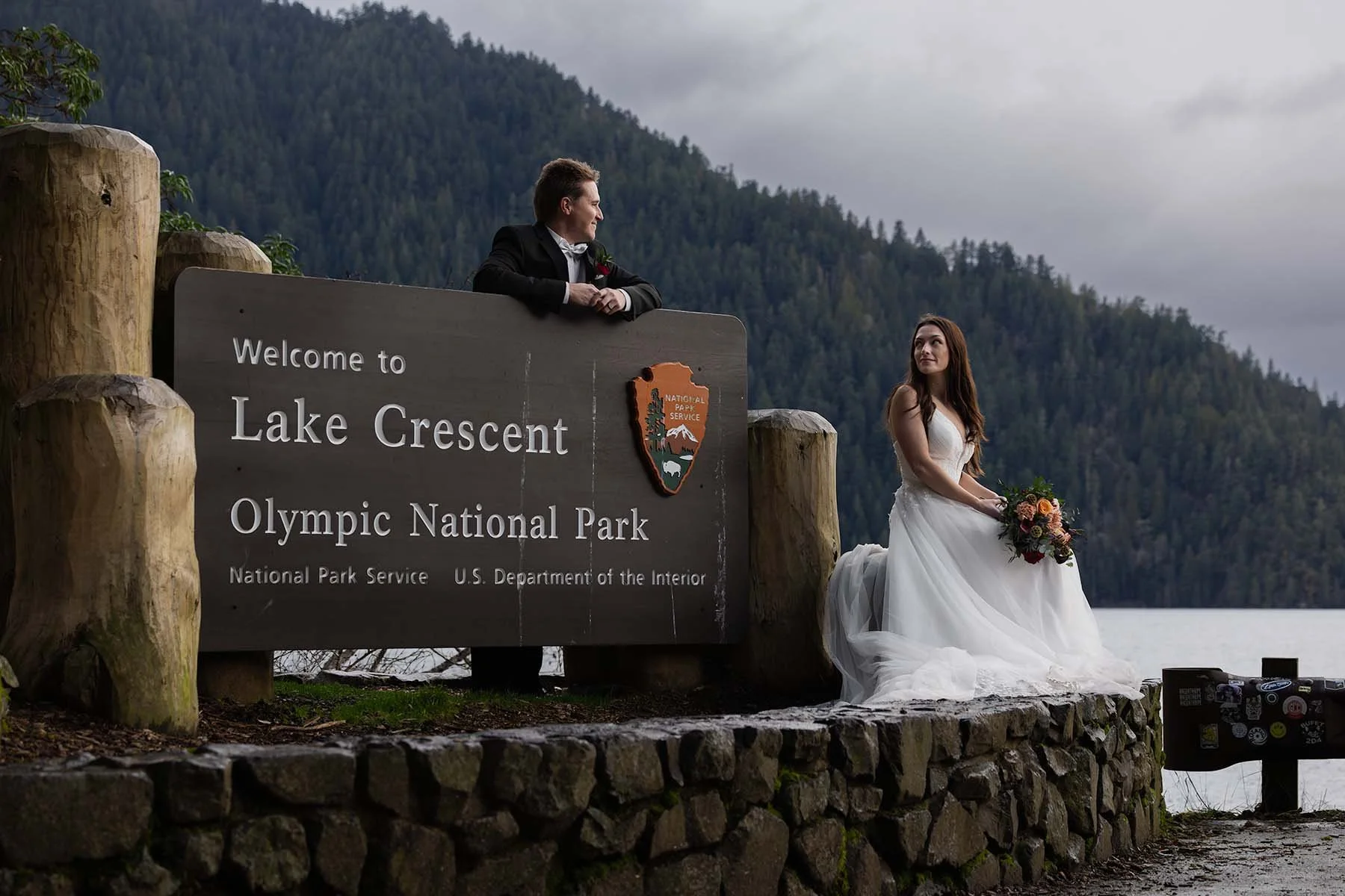 A couple in wedding attire pose with the Lake Crescent welcome sign at Olympic National Park