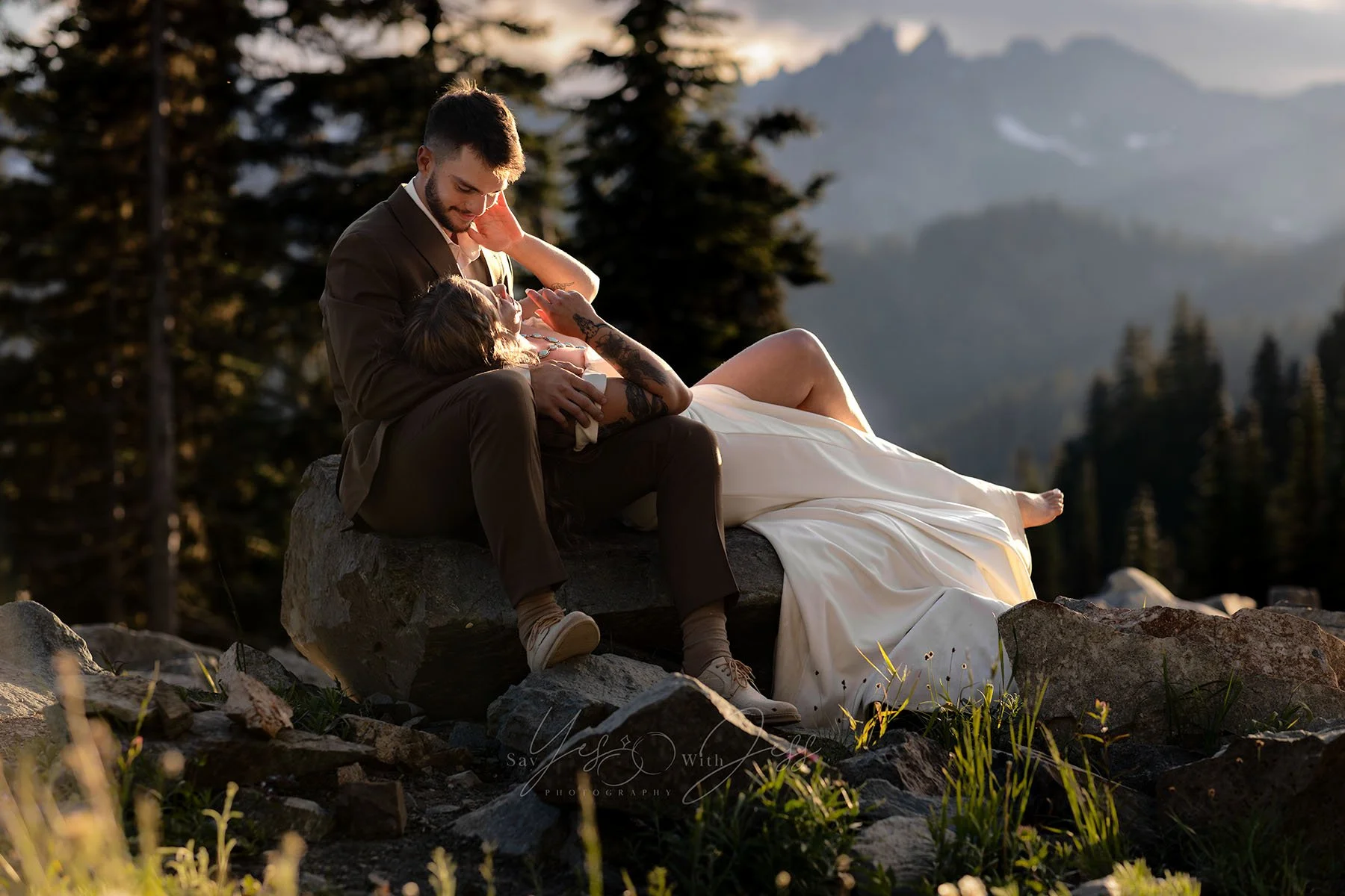 A bride lays across a flat boulder in her husband's lap and reaches for his cheek on their wedding day. They are surrounded by mountains as the sun sets.