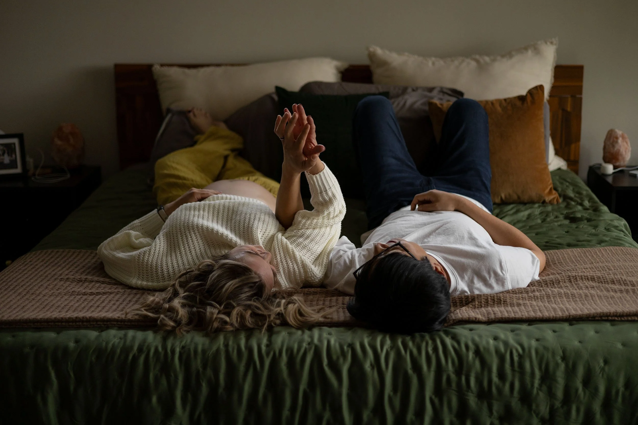 Lifestyle maternity couple lay in their bed, delicately intertwining fingers during a session in Bonney Lake, Washington.