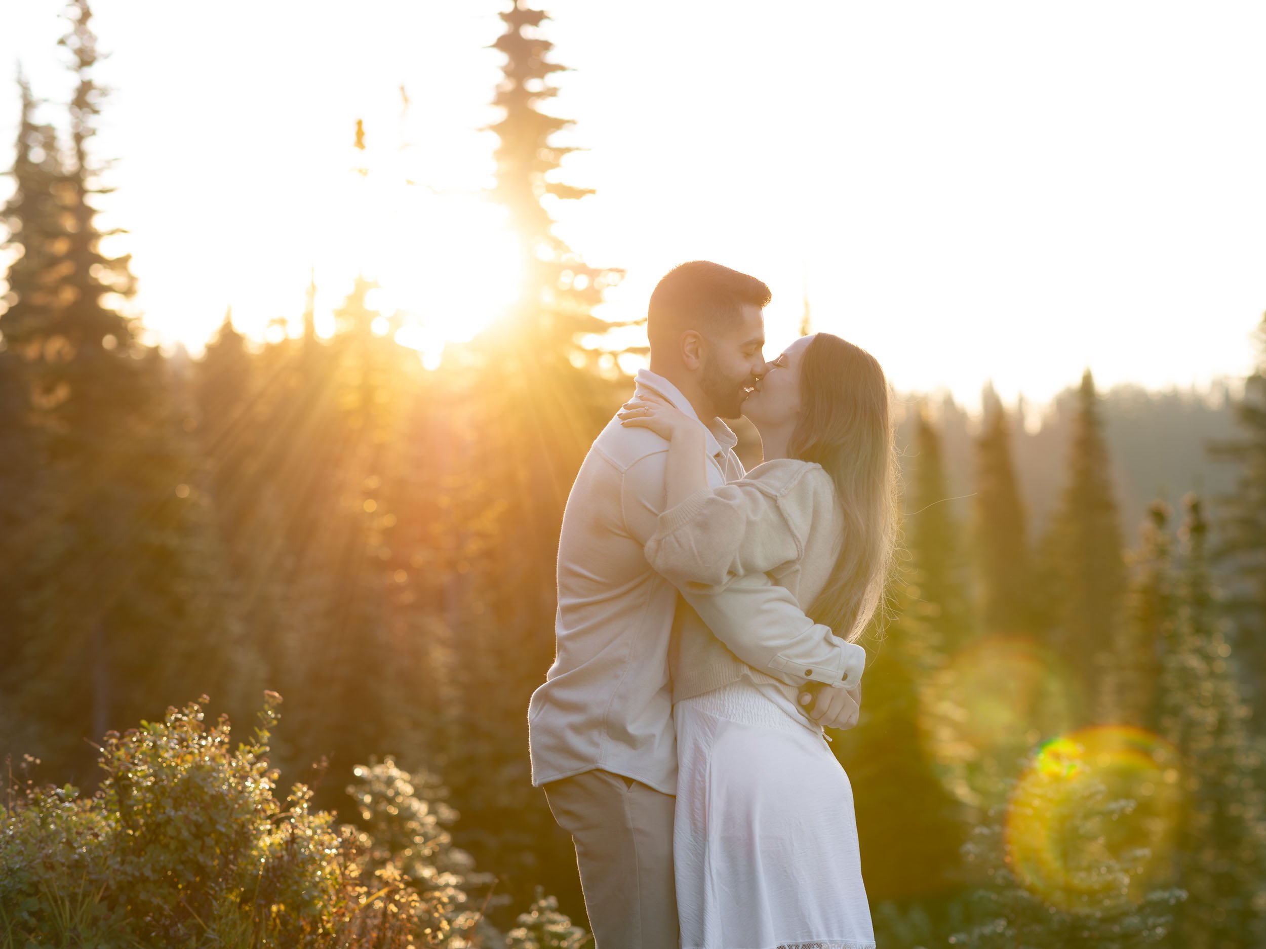 Engagement session couple share a romantic kiss at Mount Rainier National Park. The rising sun washes the scene in an orange hue and flares the lens.