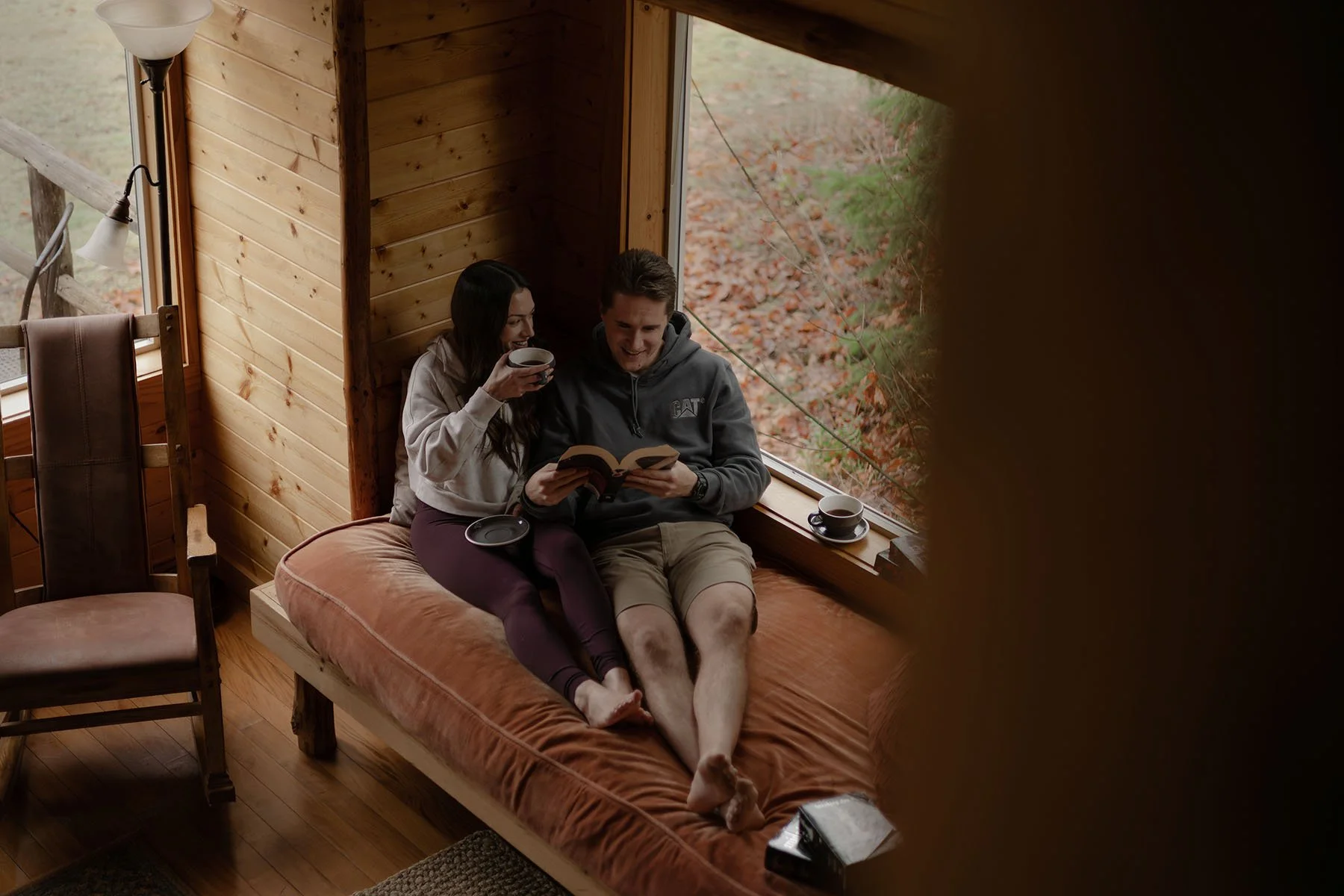 A couple snuggle up on a daybed in a cabin drinking coffee and reading a book on the morning of their elopement.