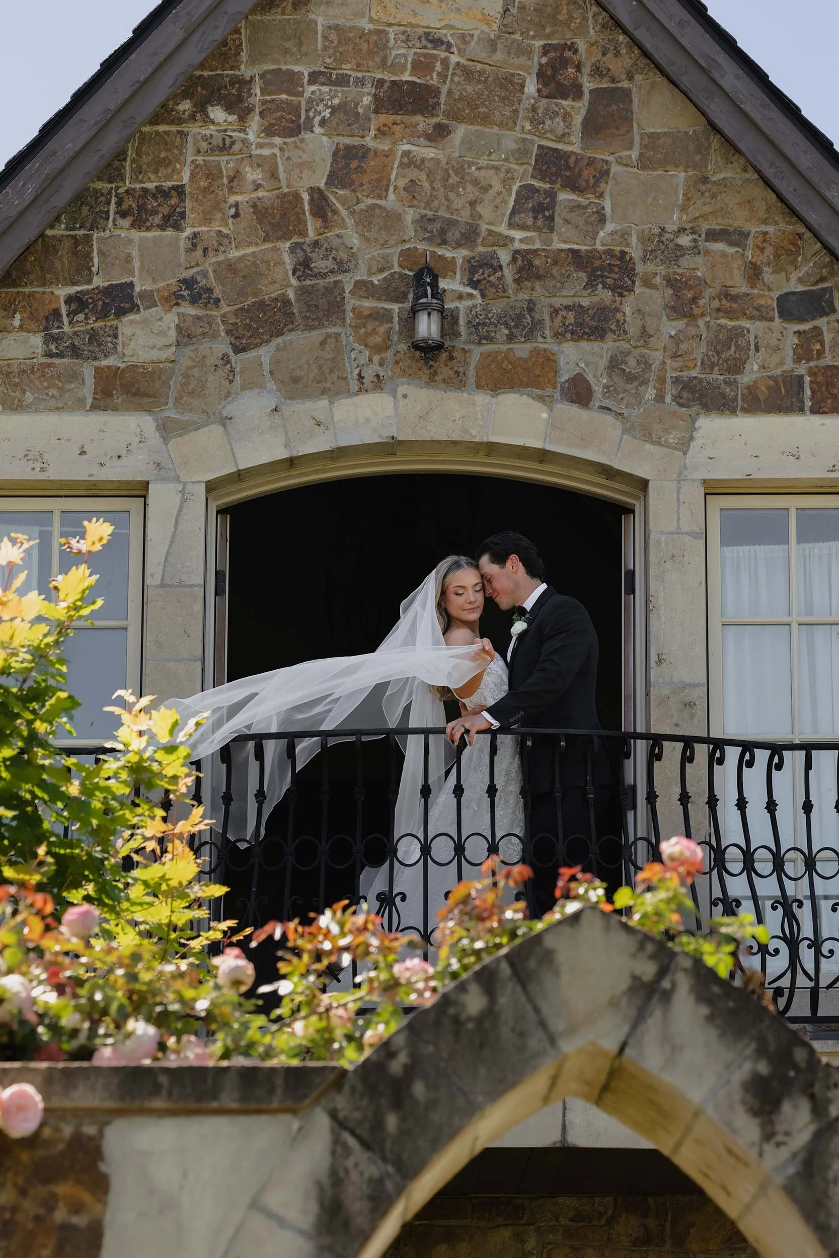A bride with a long flowing veil and her husband stand on the balcony of their European inspired wedding venue. 