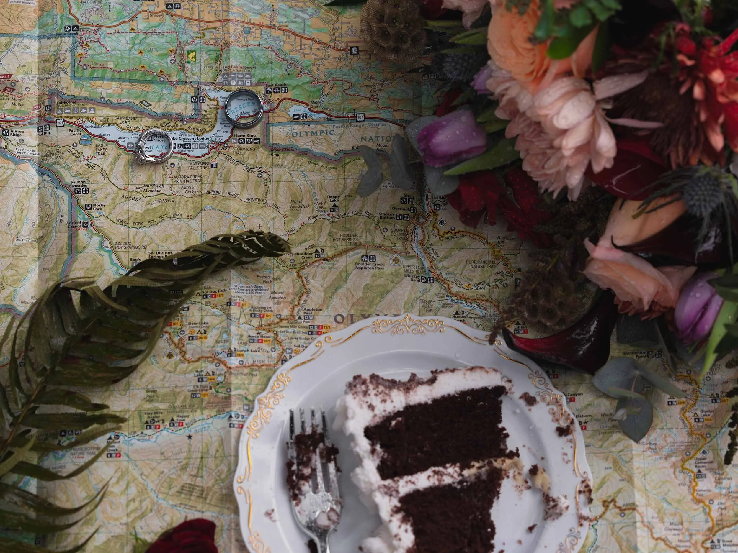 Flat lay of a map of Olympic National  Park, half-eaten wedding cake, a bridal bouquet and wedding rings positioned on Lake Crescent.