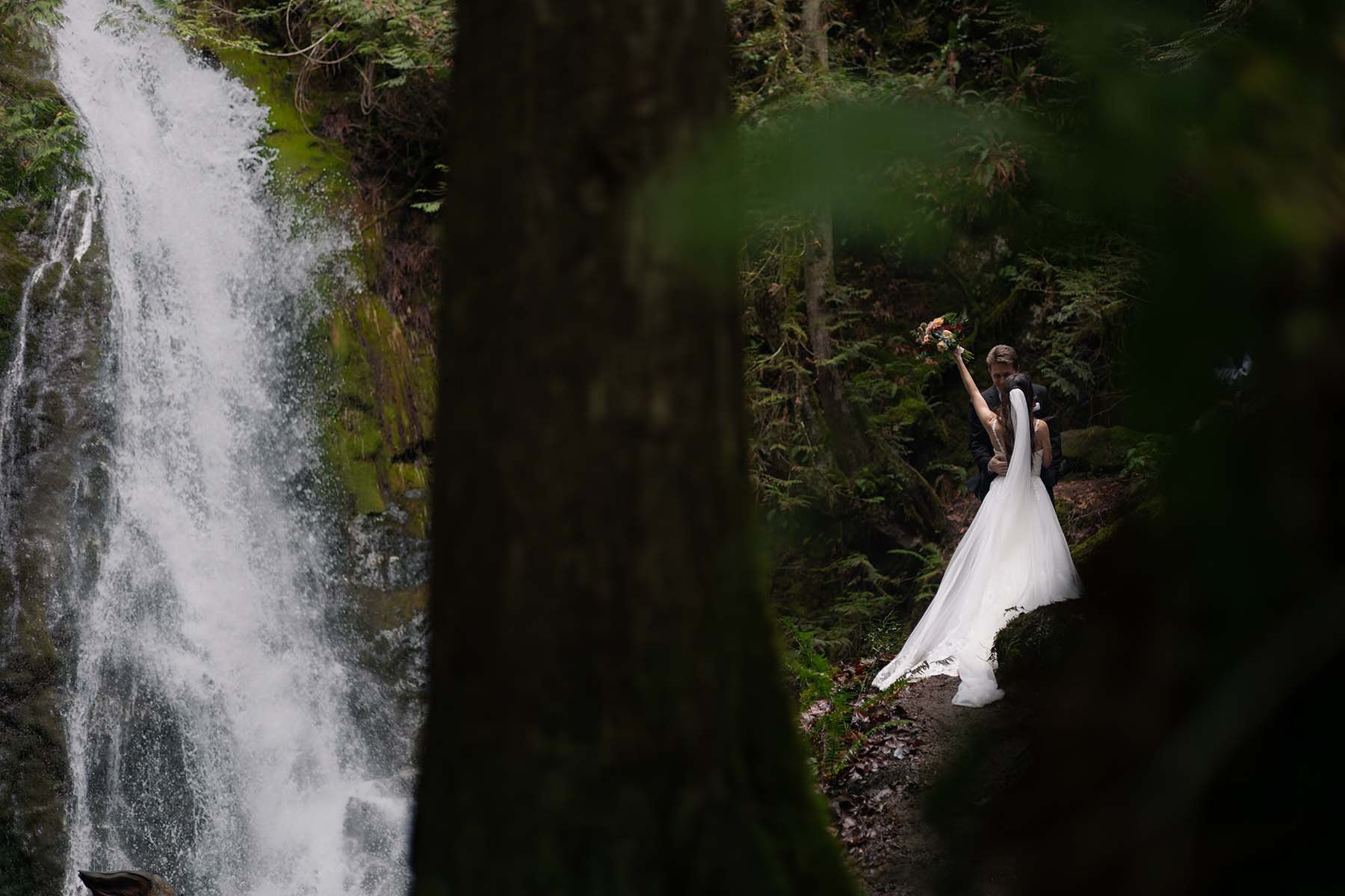 A bride throws her bouquet arm up at Madison Falls. Trees in the foreground frame the couple for dimension.