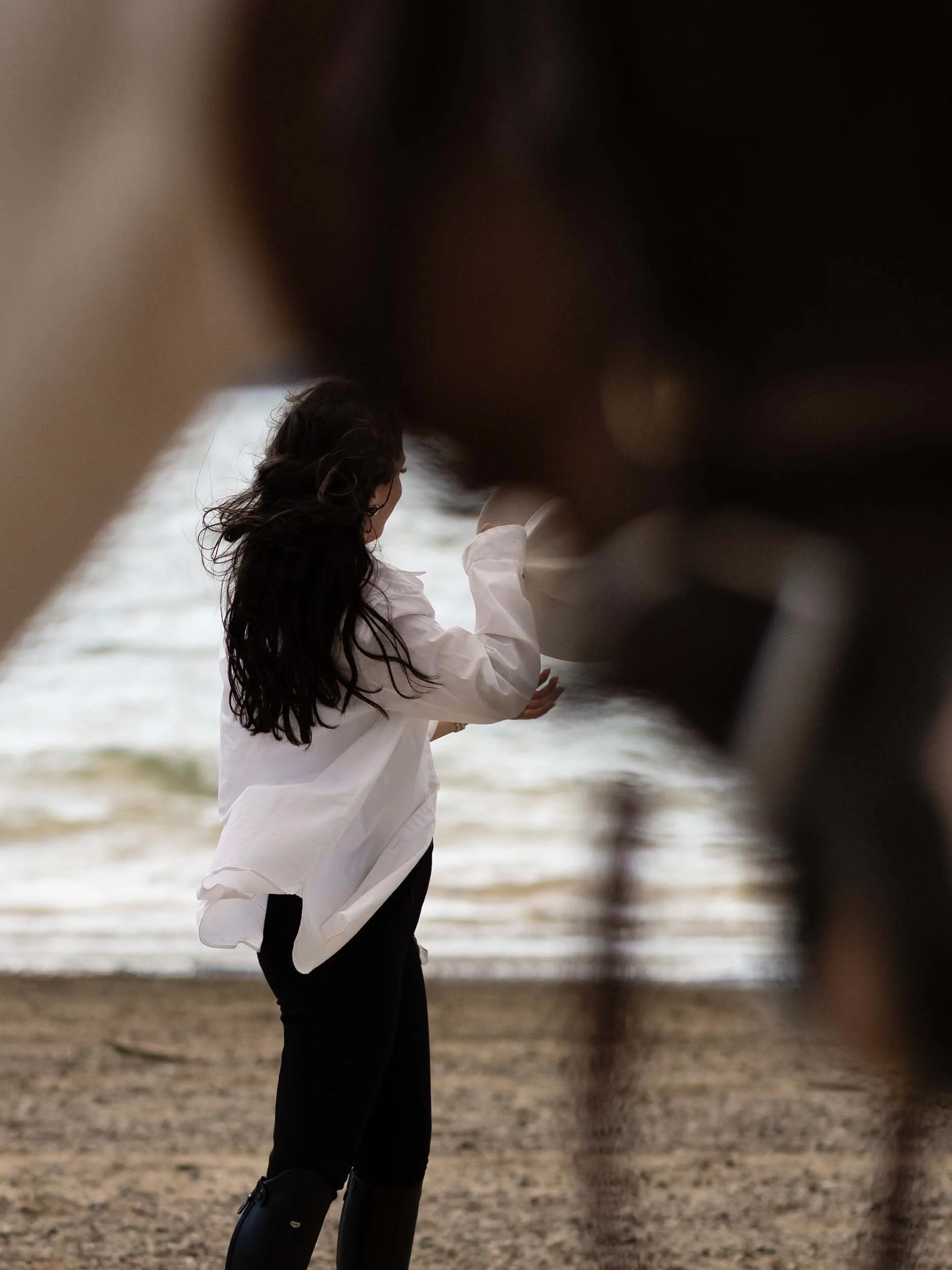 A woman in equestrian attire takes off her hat as the wind blows her hair. She is framed by a horse in the foreground
