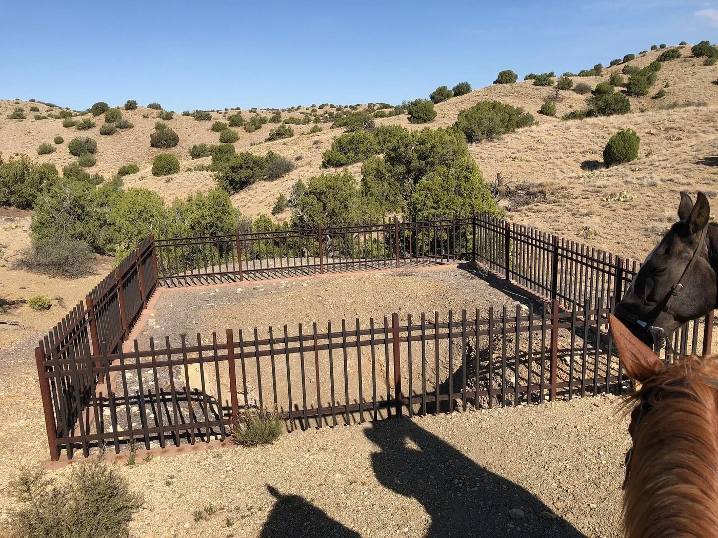 A fenced area with a saddle on the ground in a desert landscape, with a horse and part of another horse in the foreground.
