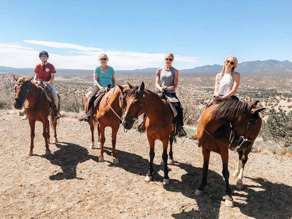 Four women riding horses in a desert landscape with mountains in the background under a blue sky.