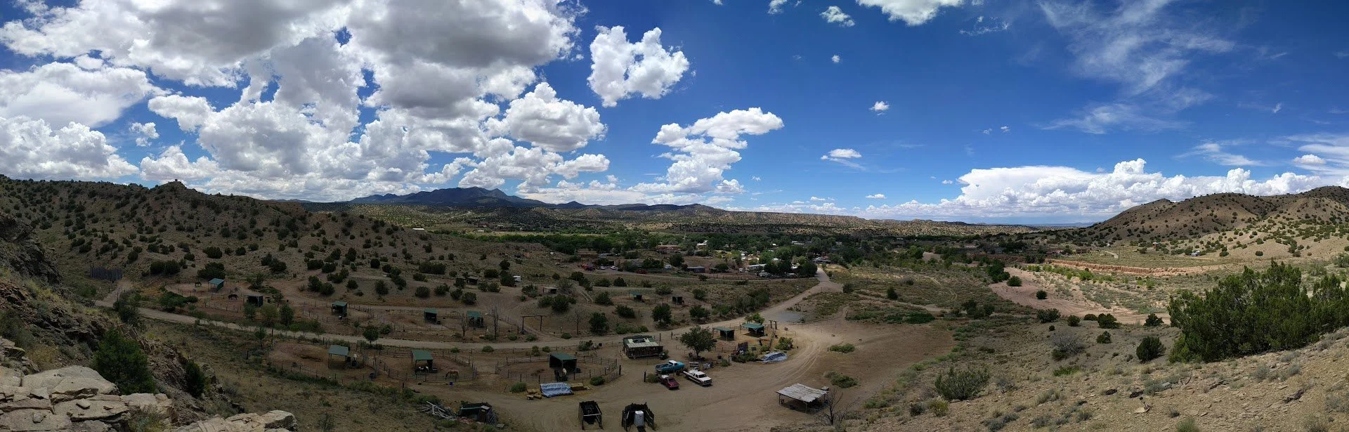 Panoramic view of a rural landscape with rolling hills, scattered trees, and a small ranch with parked vehicles, under a partly cloudy sky.