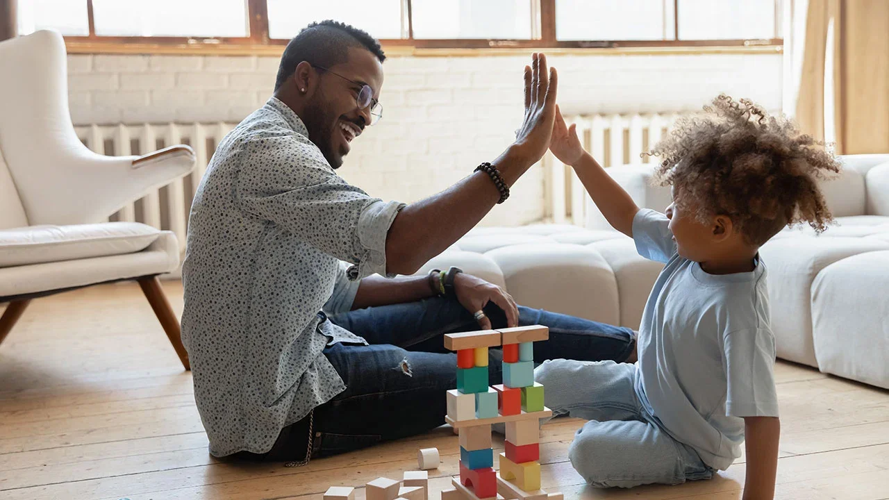 A man and a young girl are sitting on the wooden floor of a bright living room, giving each other a high five. The girl has curly hair and is wearing a light gray T-shirt and jeans, while the man has glasses and is wearing a patterned shirt and ripped jeans. There are colorful wooden blocks stacked into a tower between them.