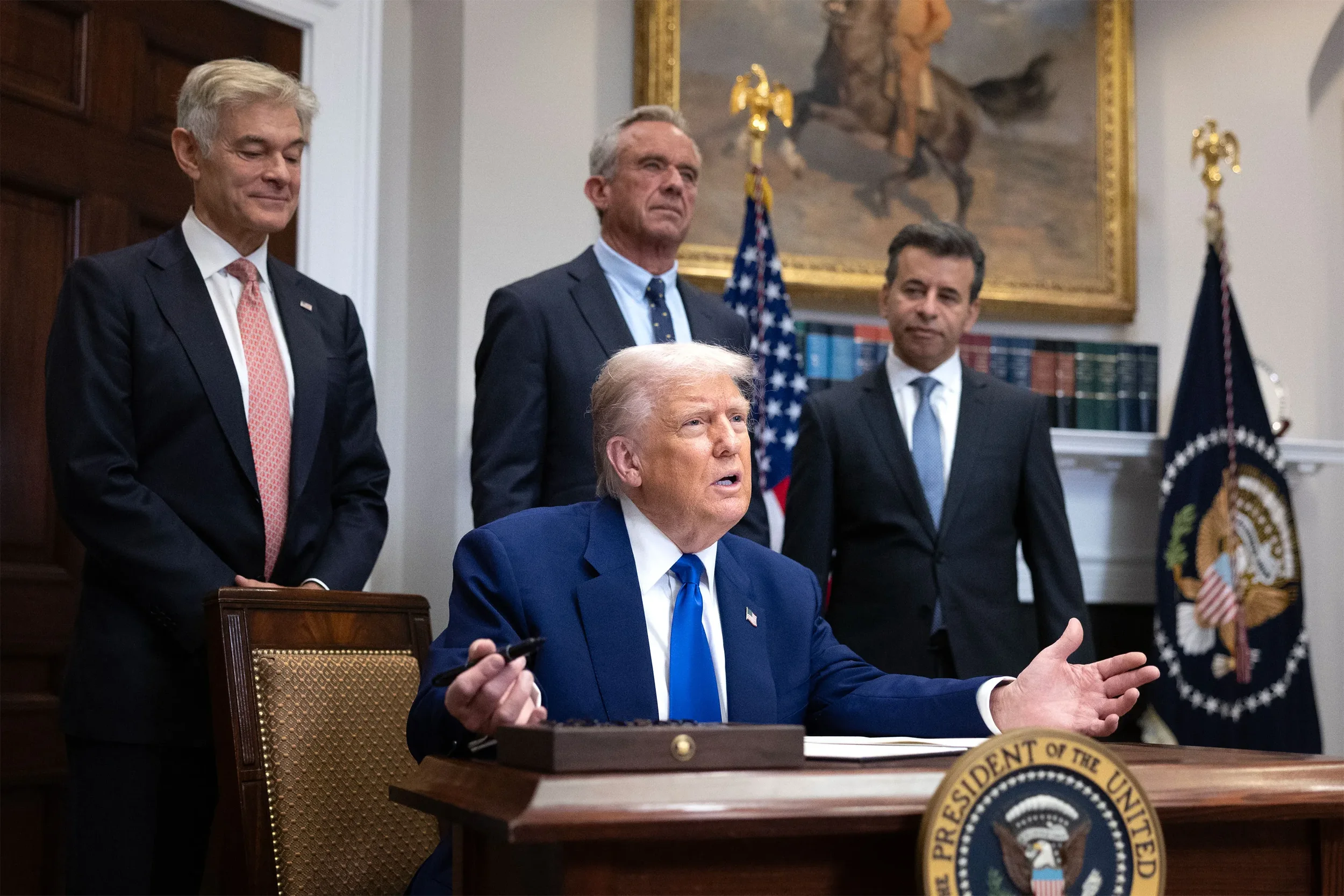 President Donald Trump seated at a desk, gesturing with his right hand, with four men standing behind him in the Oval Office. The scene includes American flags, a framed painting, and official seals.