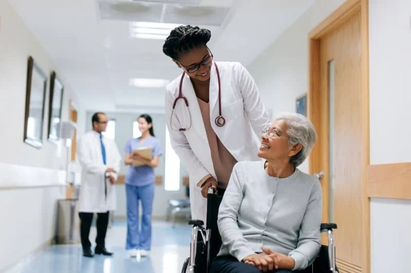A female nurse pushing an elderly woman in a wheelchair, both smiling in a hospital corridor