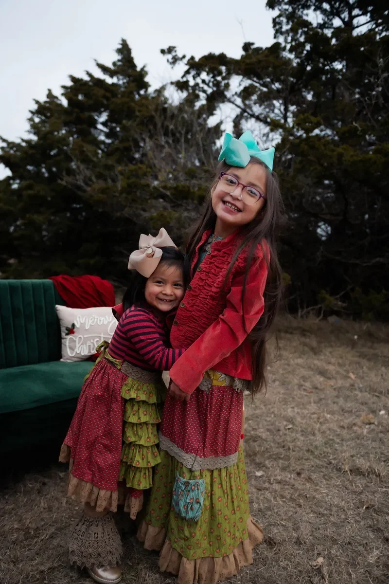 Two young girls with big bows in their hair, smiling and hugging outdoors with trees in the background. One girl is wearing a red jacket, and the other is in a striped dress. There is a green bench behind them with a pillow that says 'Merry Christmas'.
