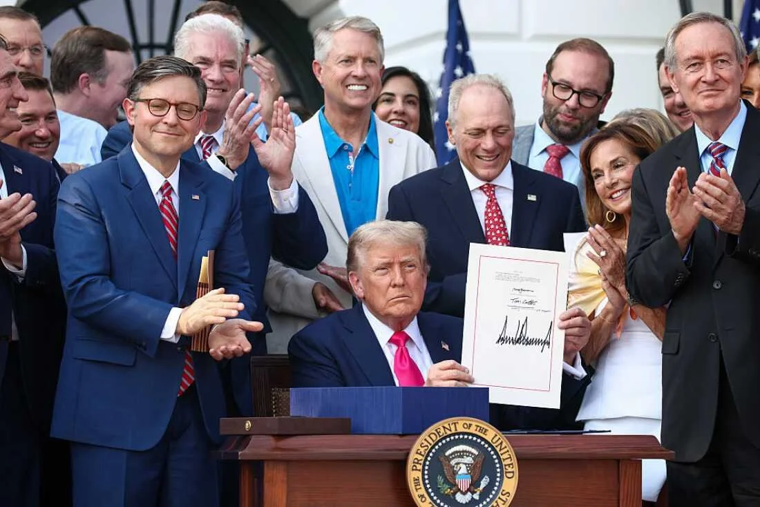 Donald Trump sitting at a desk with a presidential seal, holding a signed document, surrounded by smiling people, some clapping and others posing for a photo, outside the White House.
