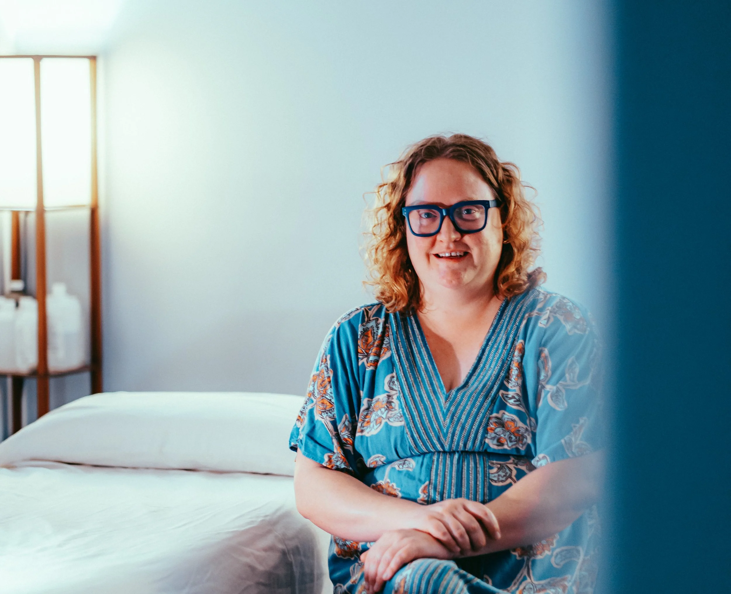 Melissa sits next to the acupuncture bed in her studio, smiling and crossing her arms on her knees. She has bright orange hair, is wearing blue glasses, and is wearing a blue dress with orange floral details.