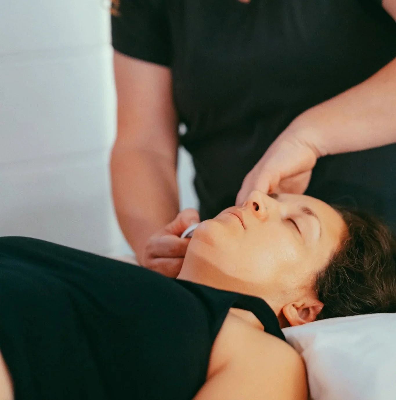 Melissa massages a client's face while they lay face up and have their eyes closed. They look deeply relaxed.