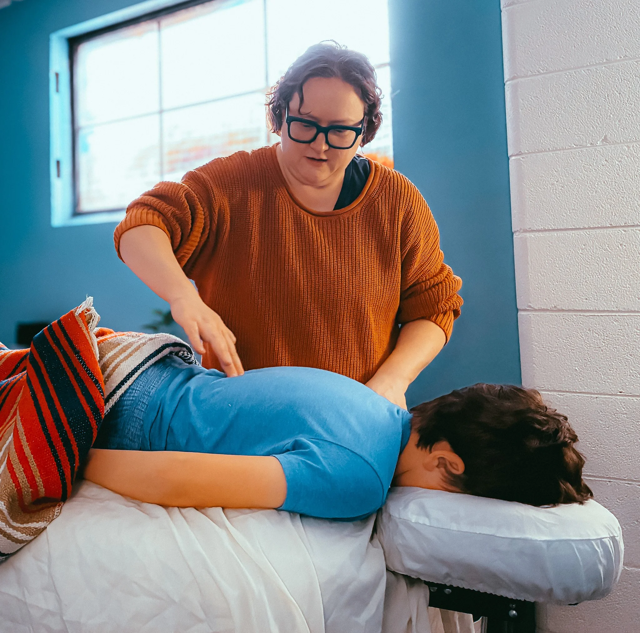 Melissa provides pediatric acupuncture care to a child laying face down on the bed. Melissa is lightly pressing two fingers into the middle of the kid's back while standing over him.
