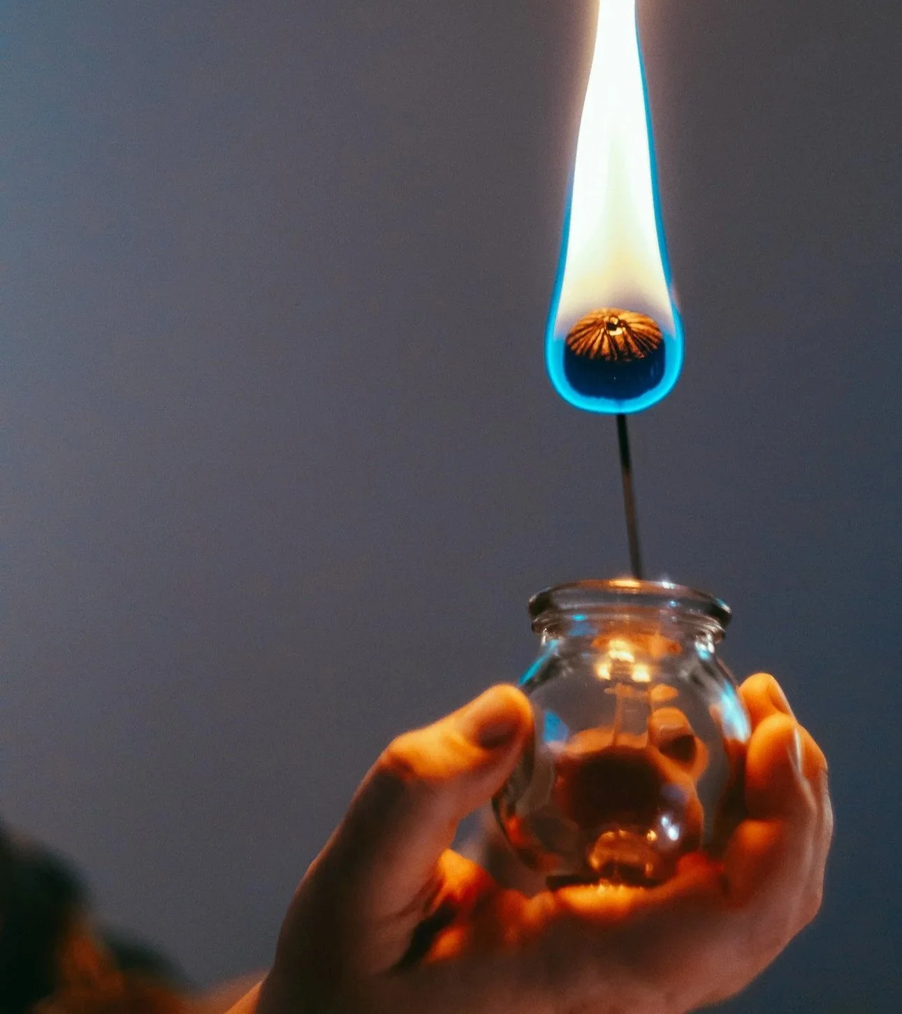 A close up image of a lit cupping wand, just being held above a glass cup in a person's hand. The background is dark grey.