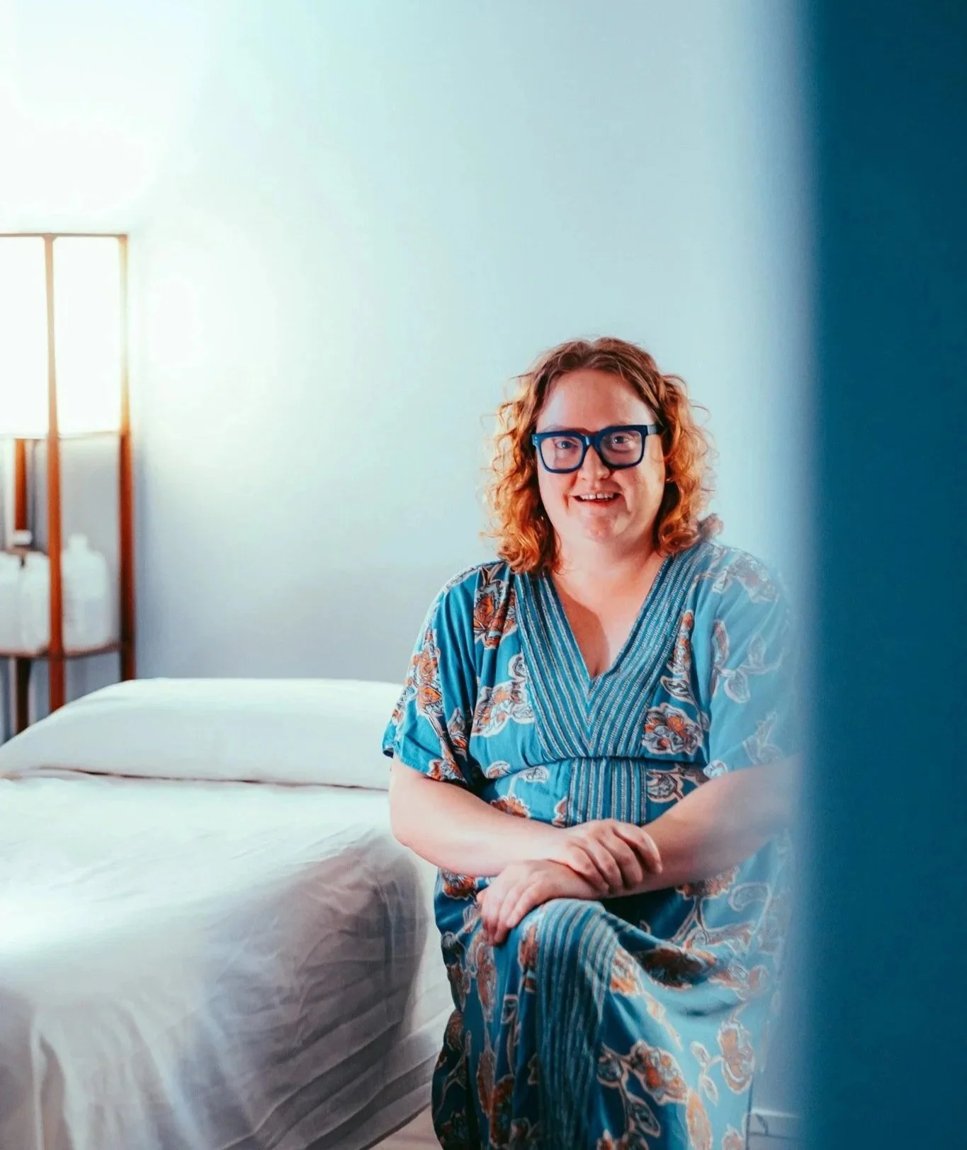 Melissa sits next to the acupuncture bed in her studio, smiling and crossing her arms on her knees. She has bright orange hair, is wearing blue glasses, and is wearing a blue dress with orange floral details.