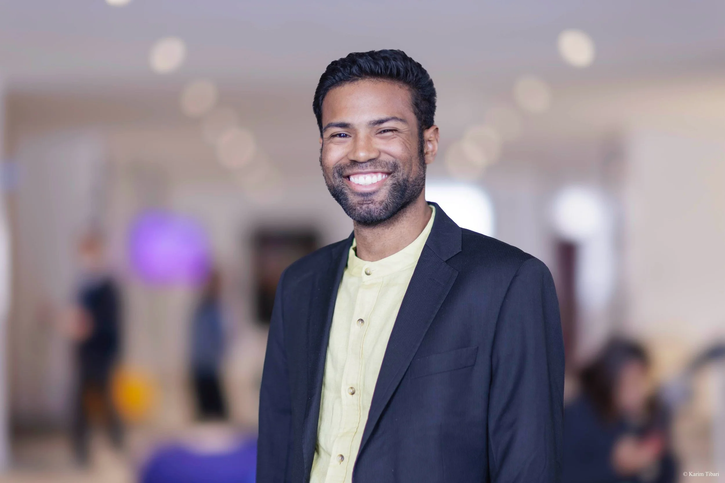 A smiling man in a dark blazer and light yellow shirt standing indoors with a blurred background of people in an office or conference setting.