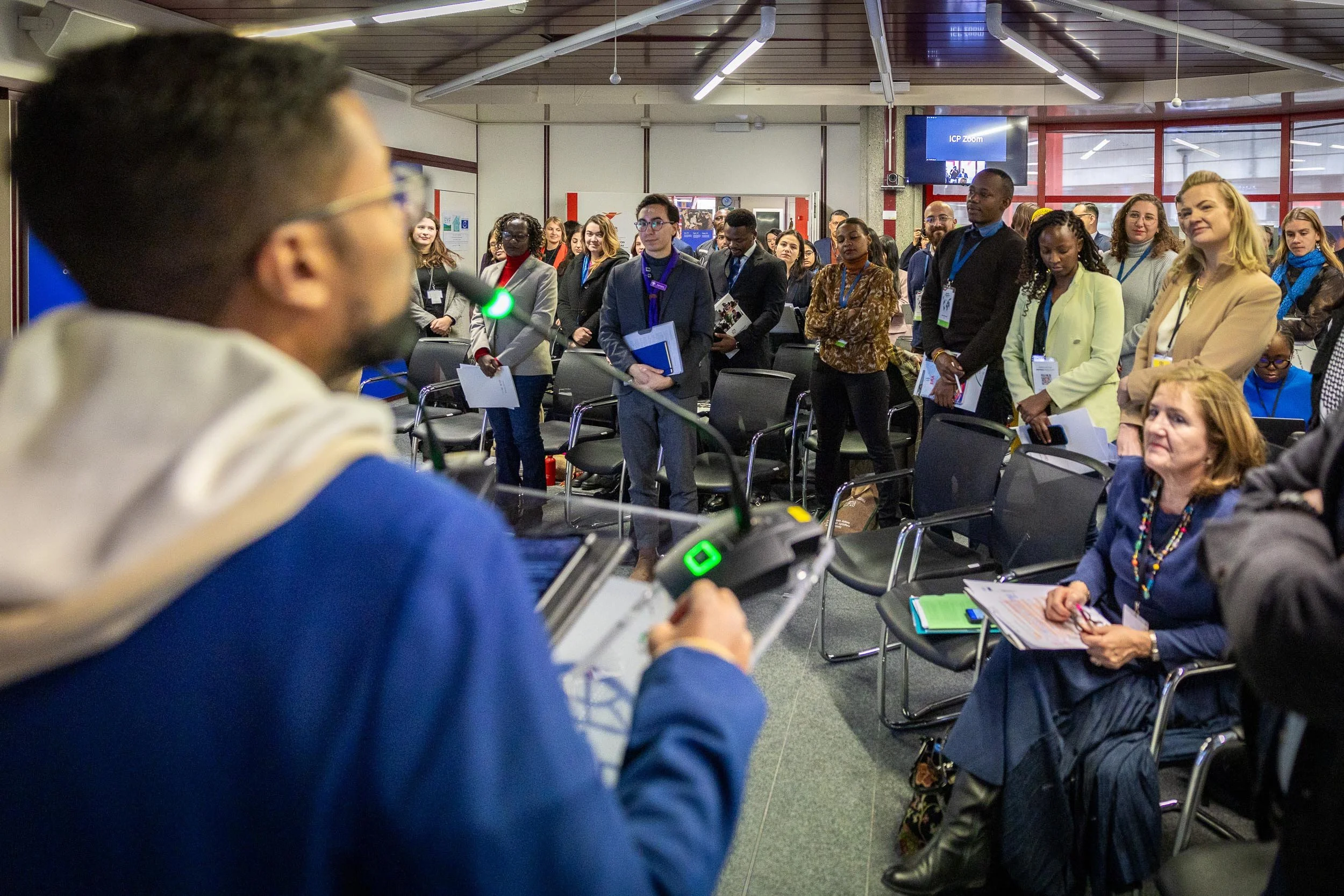 A man speaking at a conference with a microphone in front of an audience of diverse professionals standing and sitting, some holding notebooks and papers.