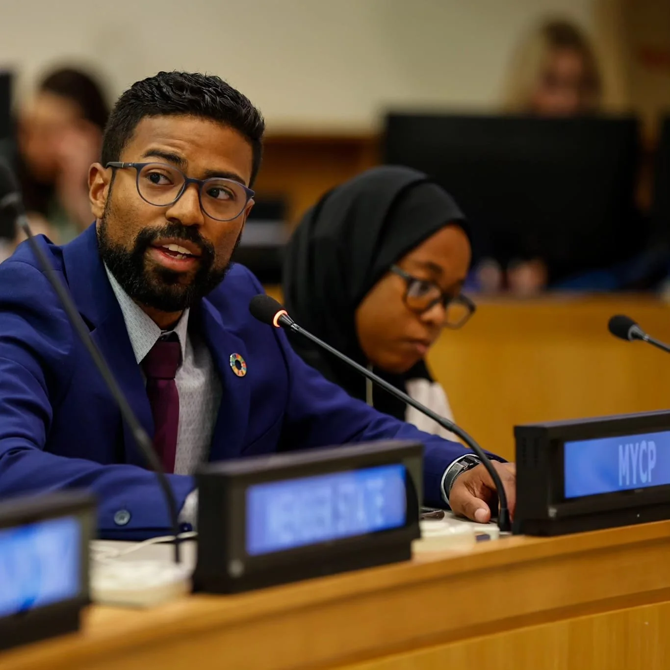 Man with glasses speaking into microphone at a conference, wearing a blue suit and a pin depicting the Sustainable Development Goals; woman beside him wearing glasses and a headscarf, looking down.