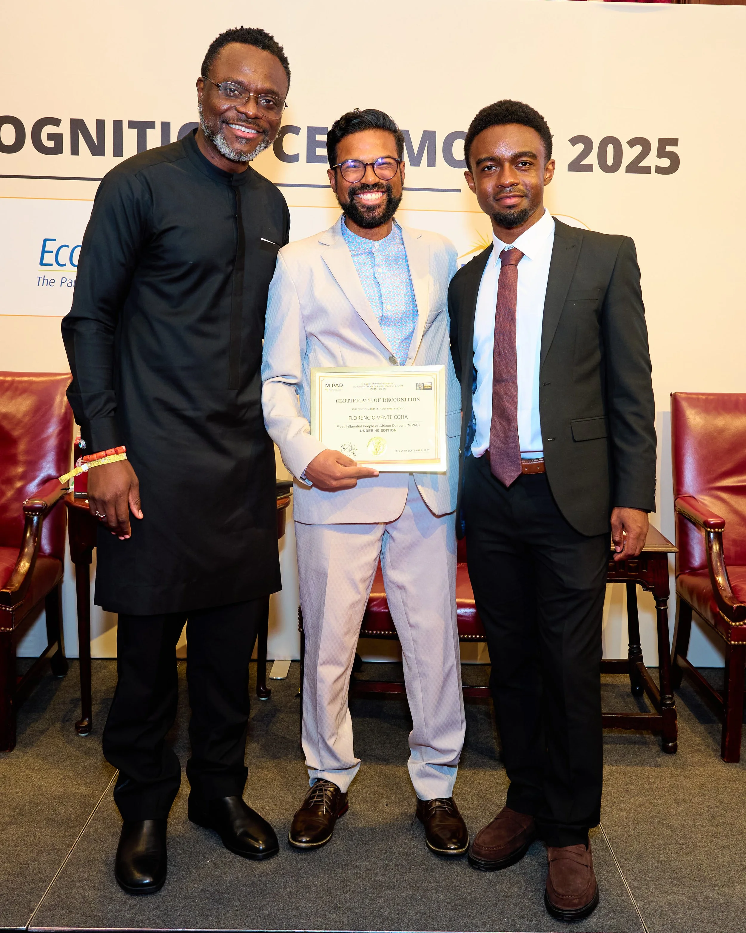 Three men in formal attire standing together at an award ceremony, with one man in the center holding a certificate. The background shows a banner with text related to the event.