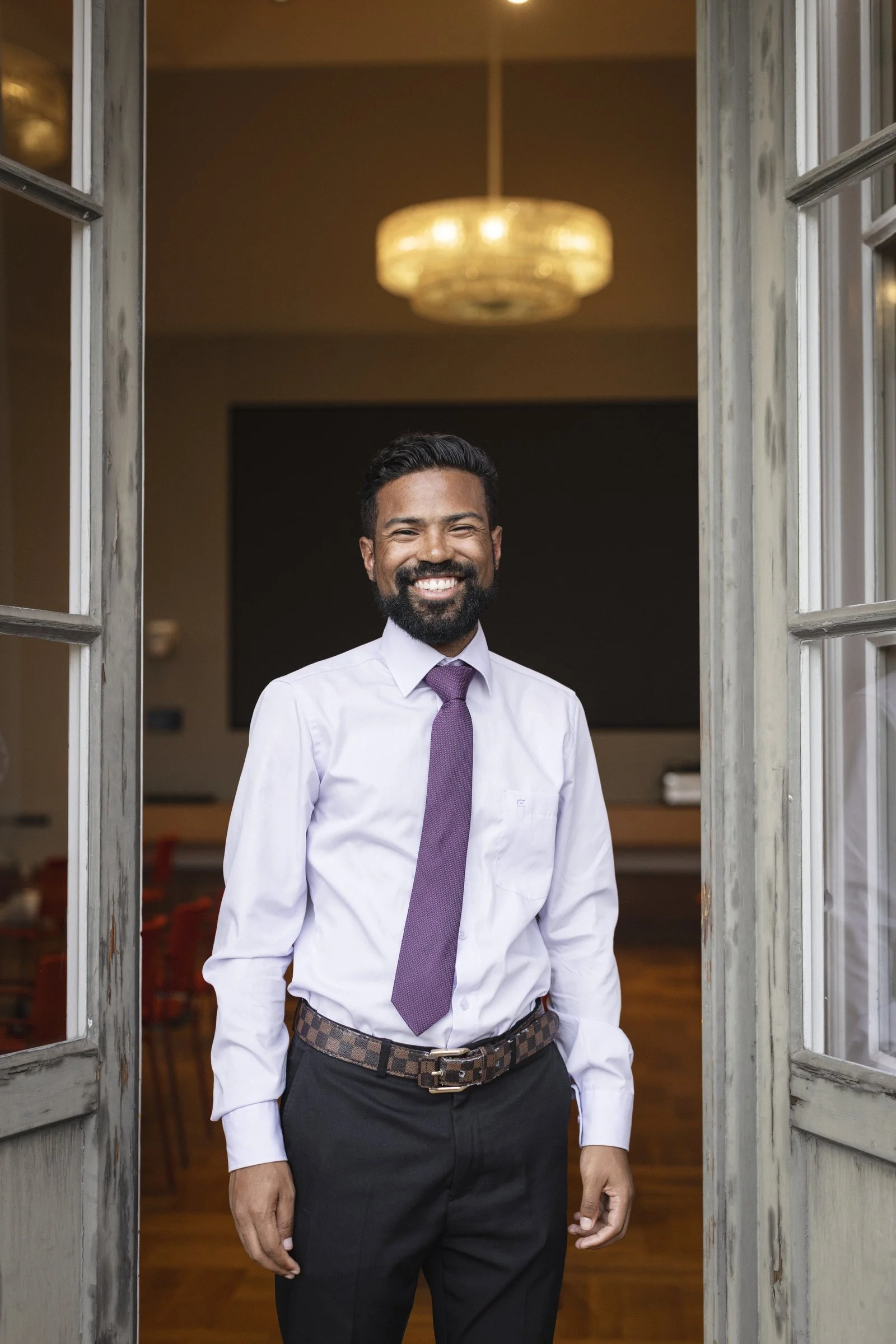 Man with dark hair and a beard smiling at the door of a room, wearing a white shirt, purple tie, and black pants with a patterned belt, inside a warmly lit conference room.