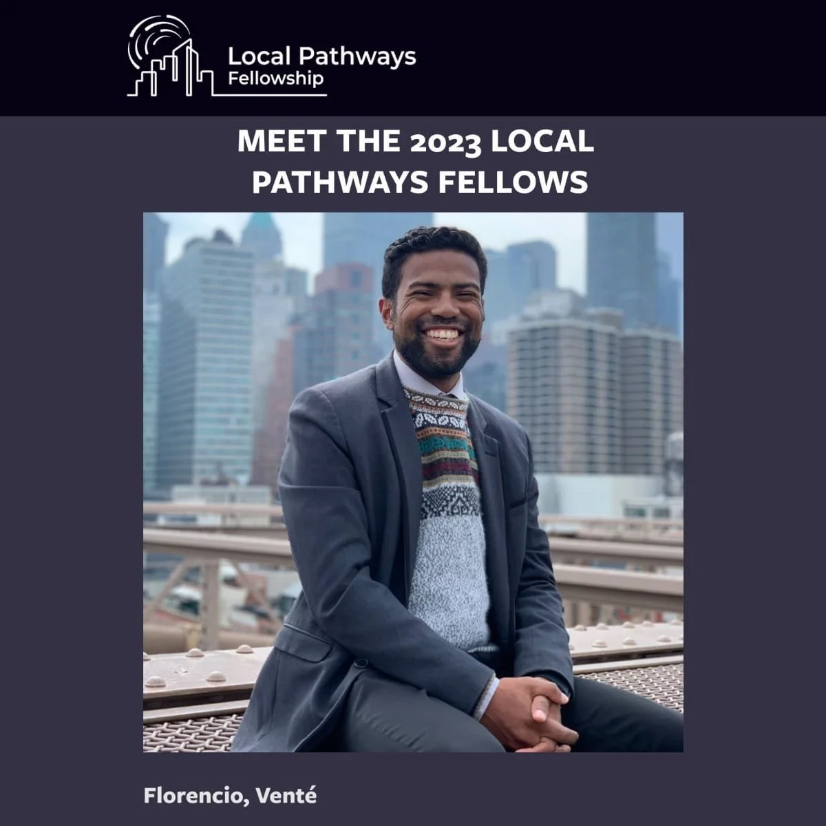 A smiling man in a suit and patterned sweater sitting outdoors with city skyscrapers in the background.