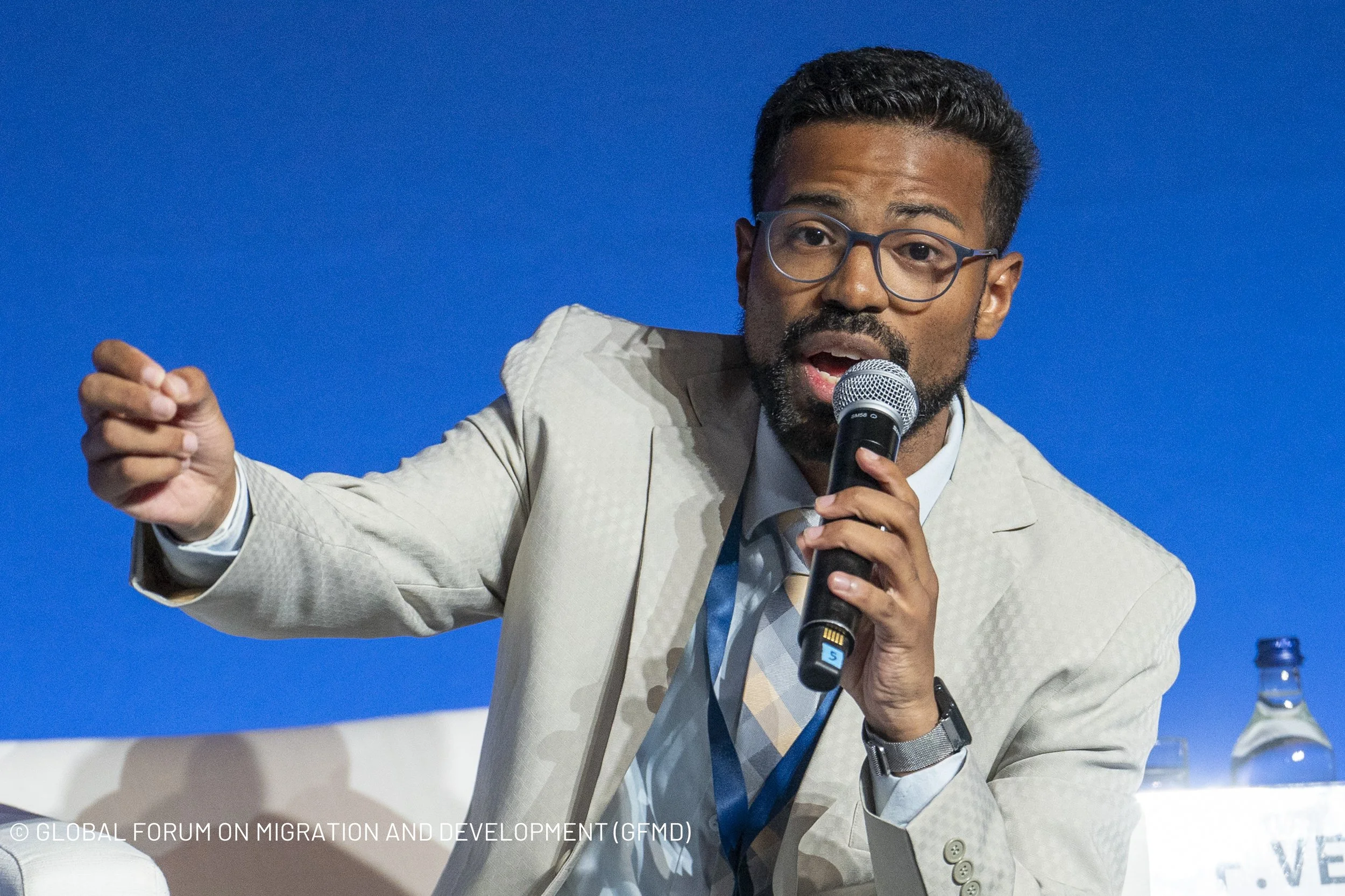 A man with glasses and a beard, wearing a light-colored suit and tie, speaking into a microphone at a conference with a blue background.