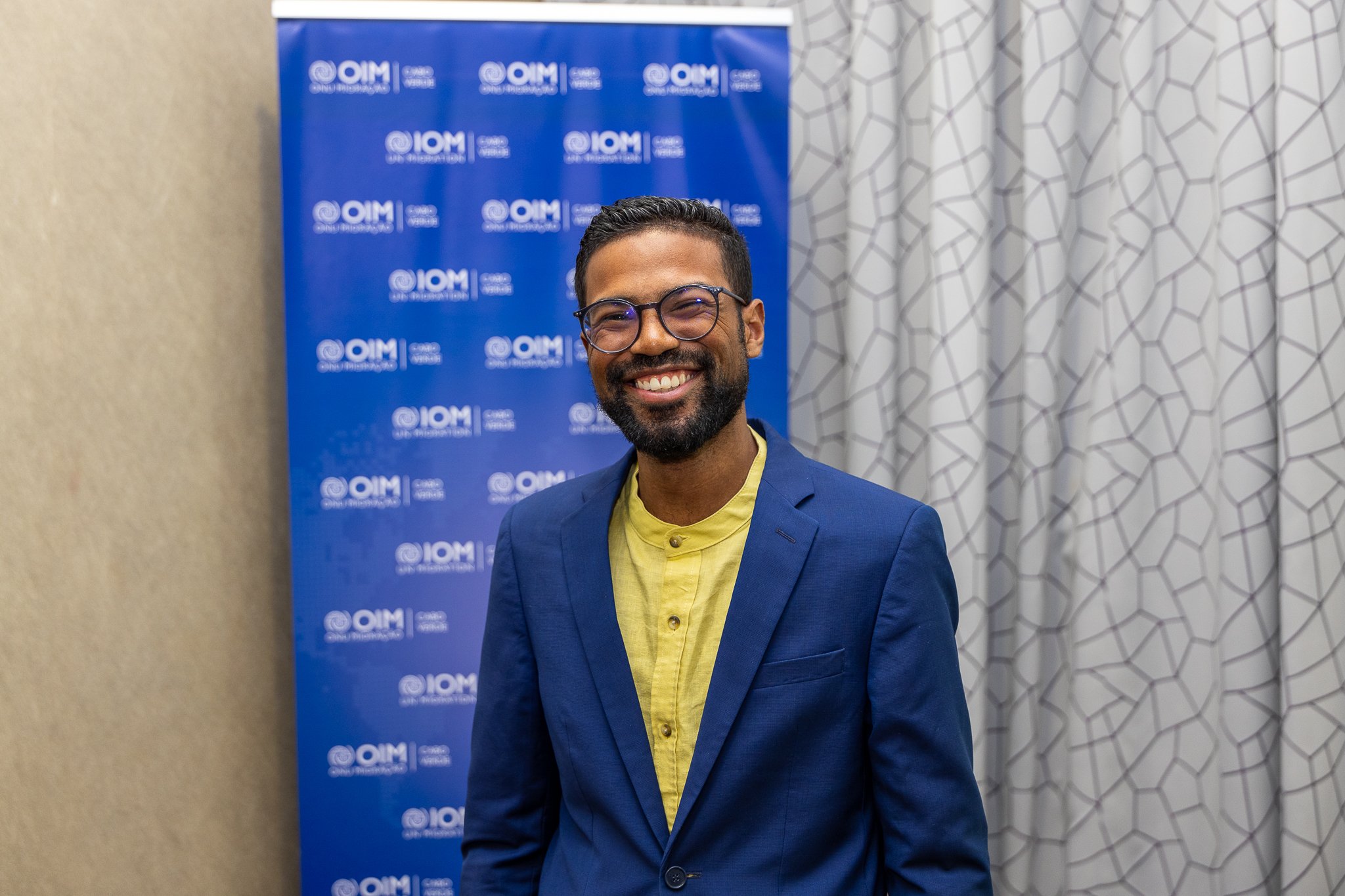 Smiling man wearing glasses, a green shirt, and a blue blazer in front of a blue backdrop with the QIM logo and a geometric-patterned curtain behind him.