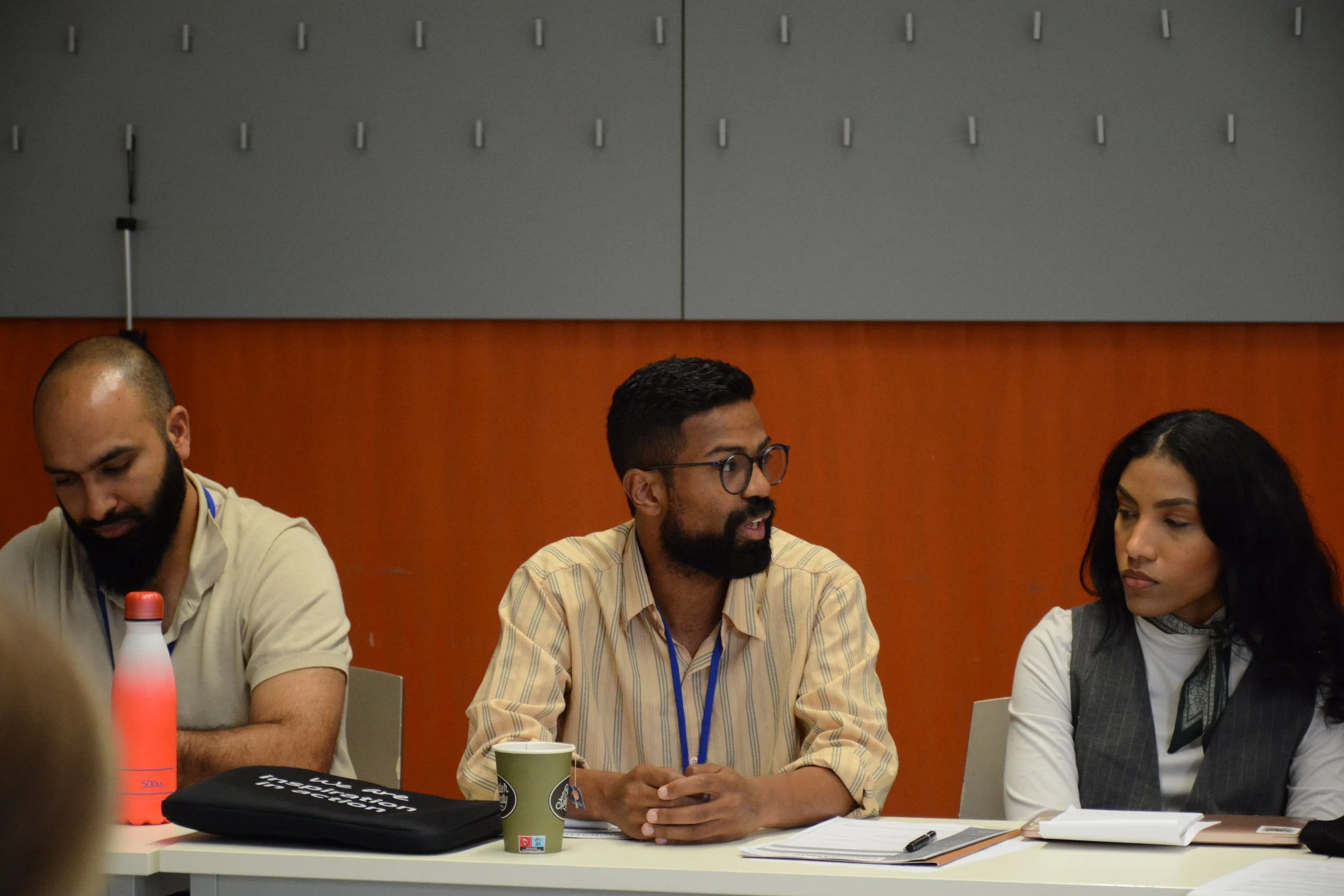 People sitting at a table in a conference room, engaged in discussion. One man with glasses and a beard is speaking, while a woman on his right and another man on his left are listening.