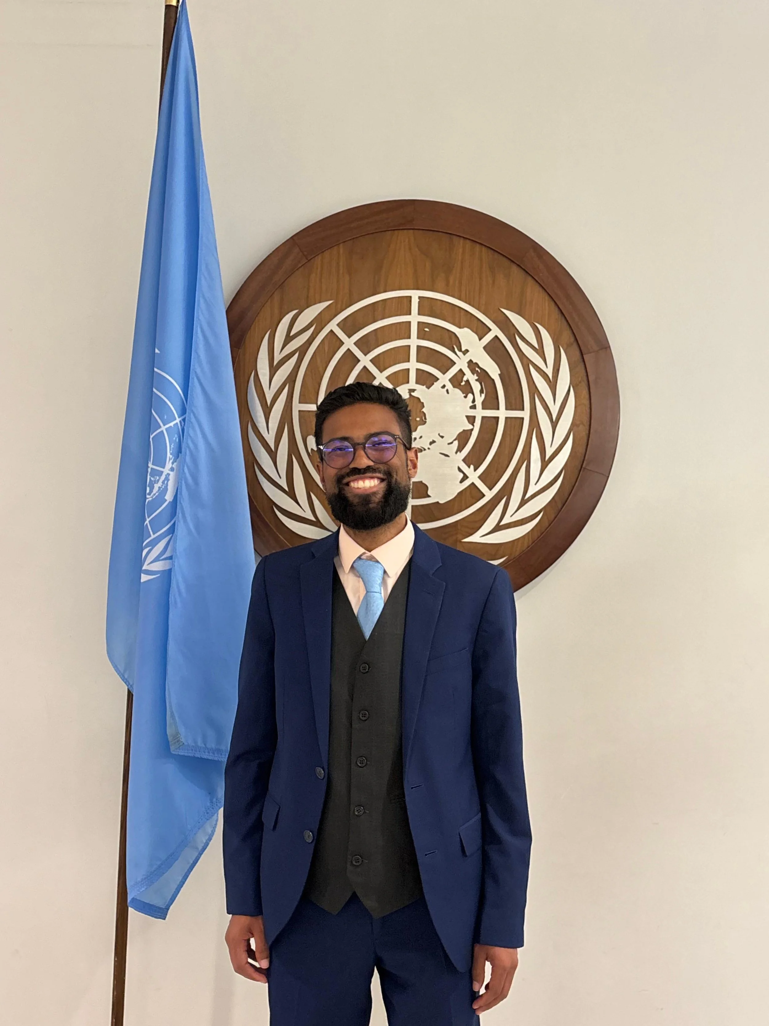 Florencio dressed in a blue suit standing in front of a United Nations emblem, with a United Nations flag to his left.