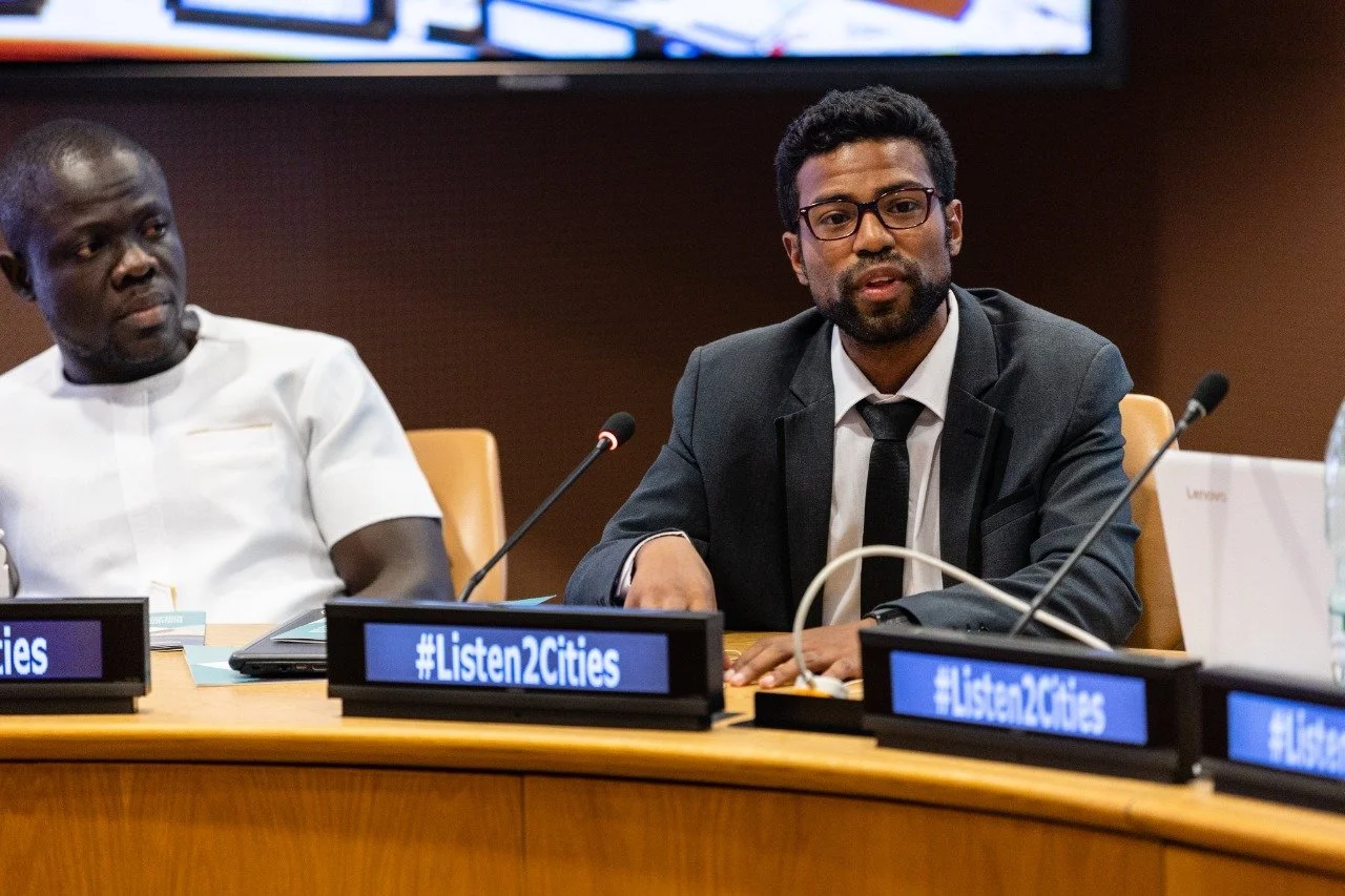Two men sitting at a conference table with microphones, one in a white shirt, the other in a dark suit and glasses, in a professional setting with #Listen2Cities displayed on the screens in front of them.