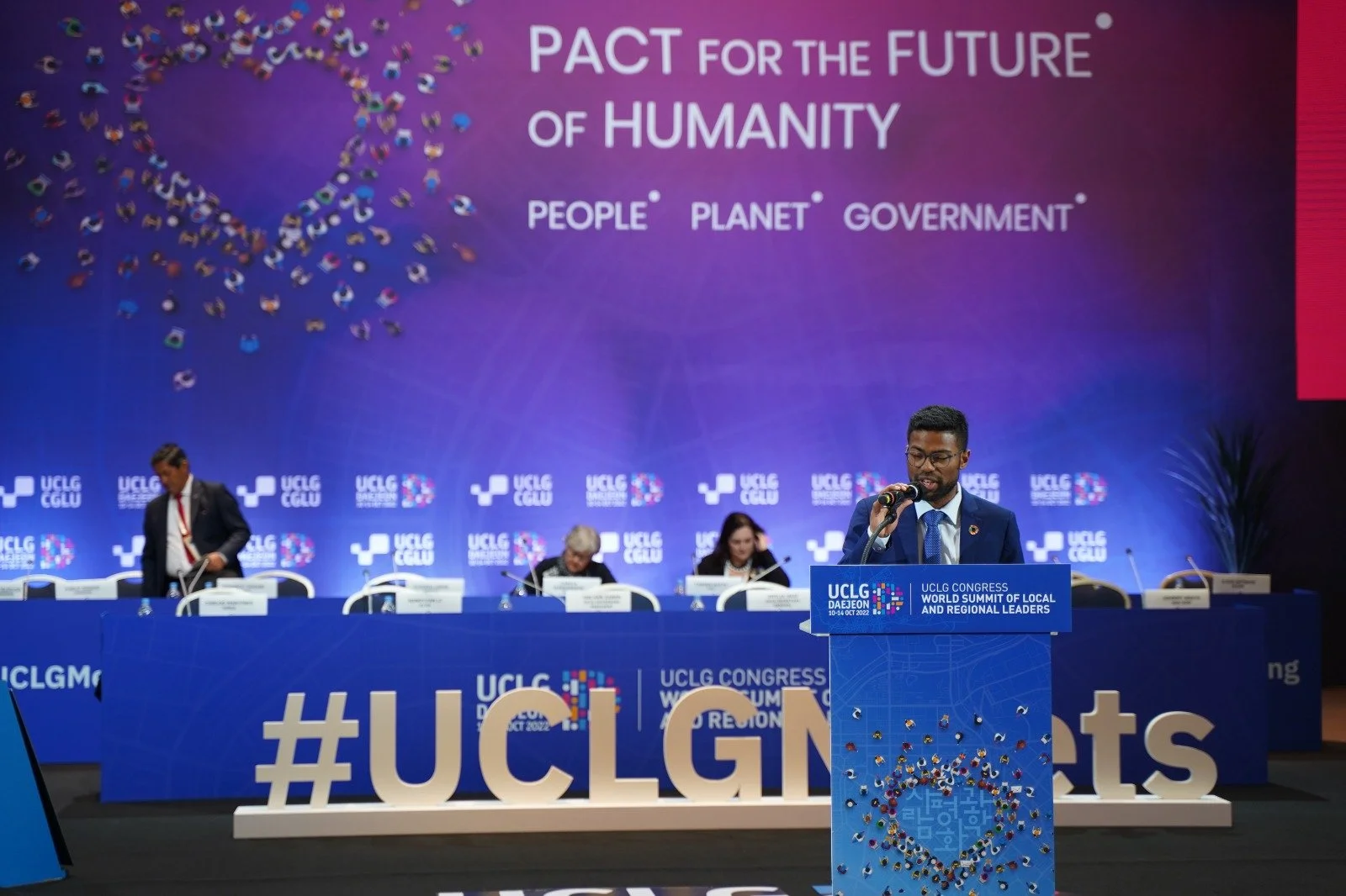 A man speaking at a podium during a conference with a blue backdrop and several people seated at a long table behind him. The backdrop features a heart shape made of small flags, the text 'PACT FOR THE FUTURE OF HUMANITY,' and words 'PEOPLE,' 'PLANET