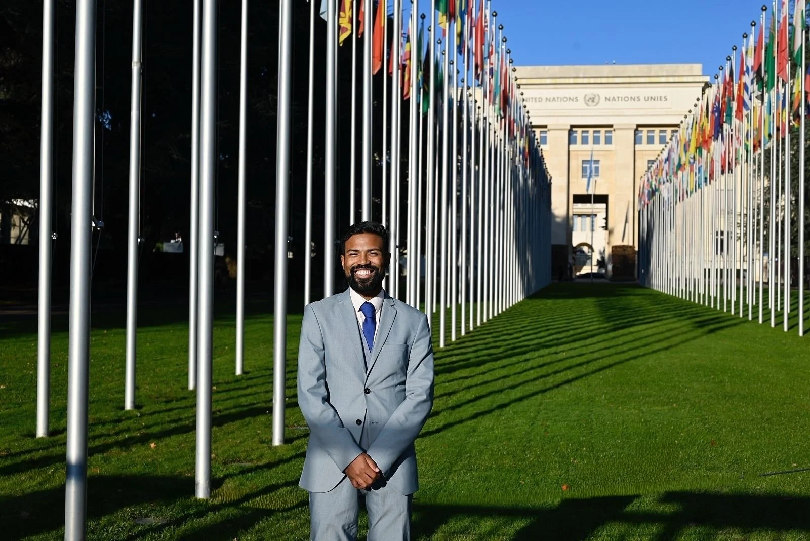 A man in a light gray suit stands smiling in front of the United Nations building, with flagpoles and flags lining both sides of a path leading to the entrance on a sunny day.