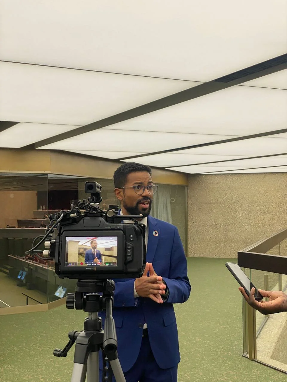 Man in blue suit being interviewed with camera on tripod and person holding a smartphone.