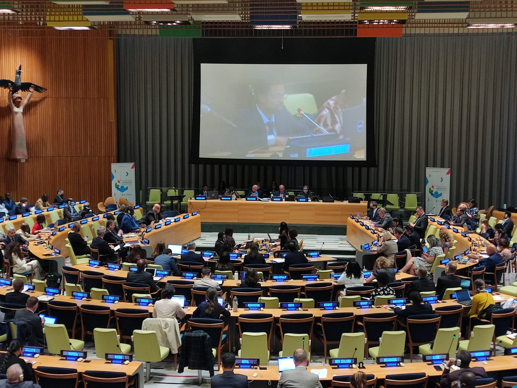 United Nations conference room with delegates seated around curved tables, microphones, nameplates, and laptops, a large screen displaying a speaker, and a sculpture of a woman with wings on the wall.